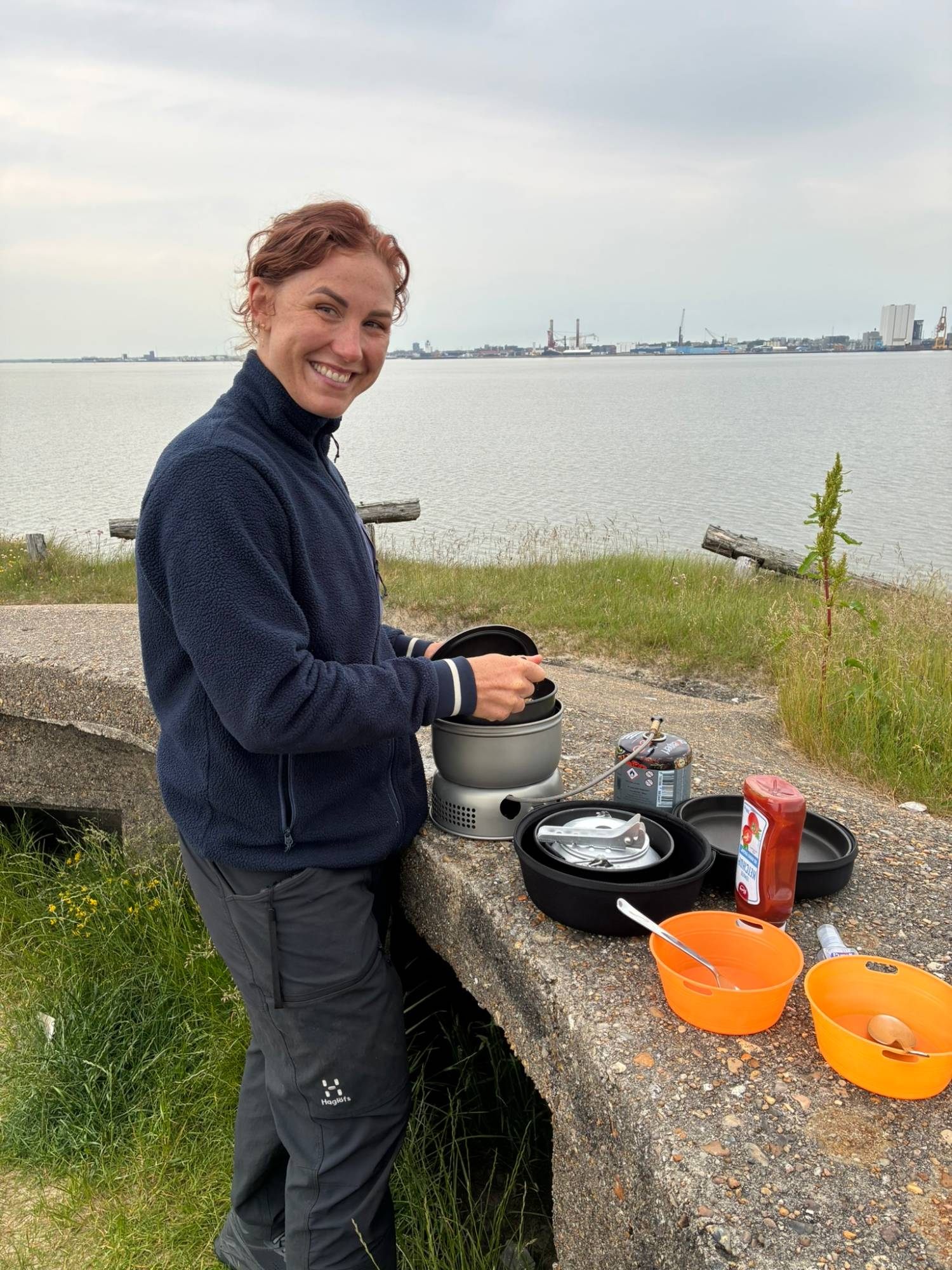 Kristine smiling while cooking on a portable camping stove on top of a concrete bunker, with Esbjerg harbour across the water