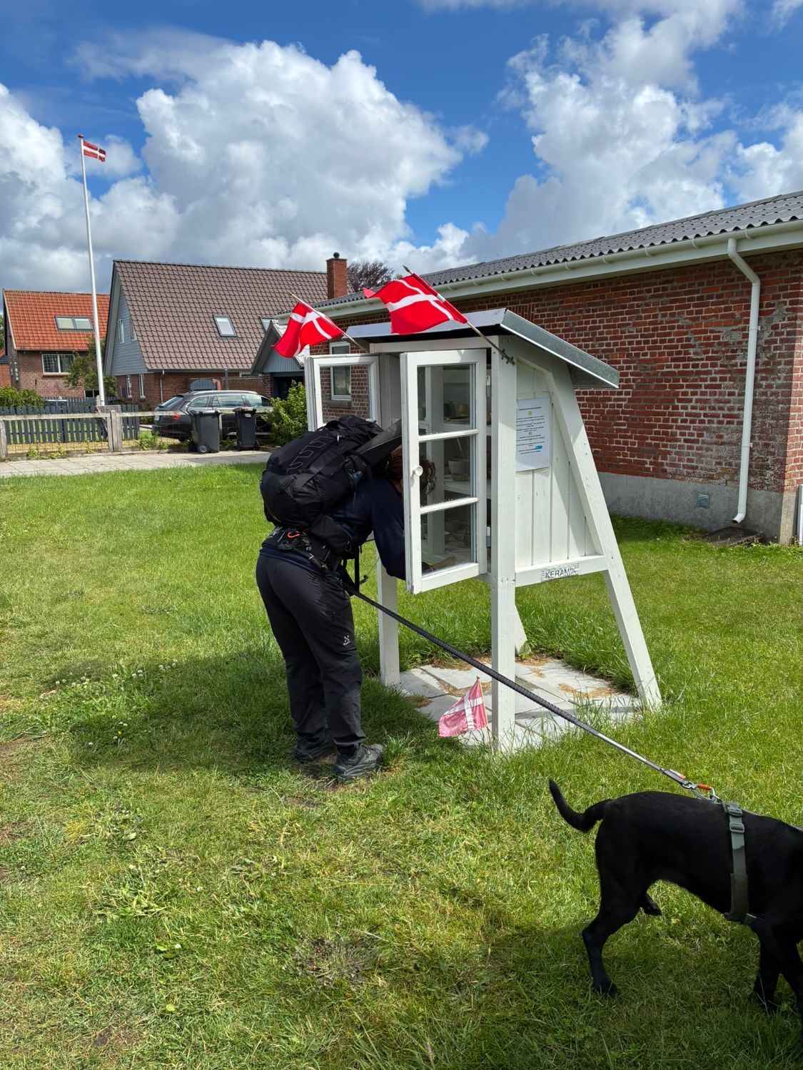 Kristine with a backpack peering into a small white roadside ceramics cabinet decorated with Danish flags, Maggie beside her