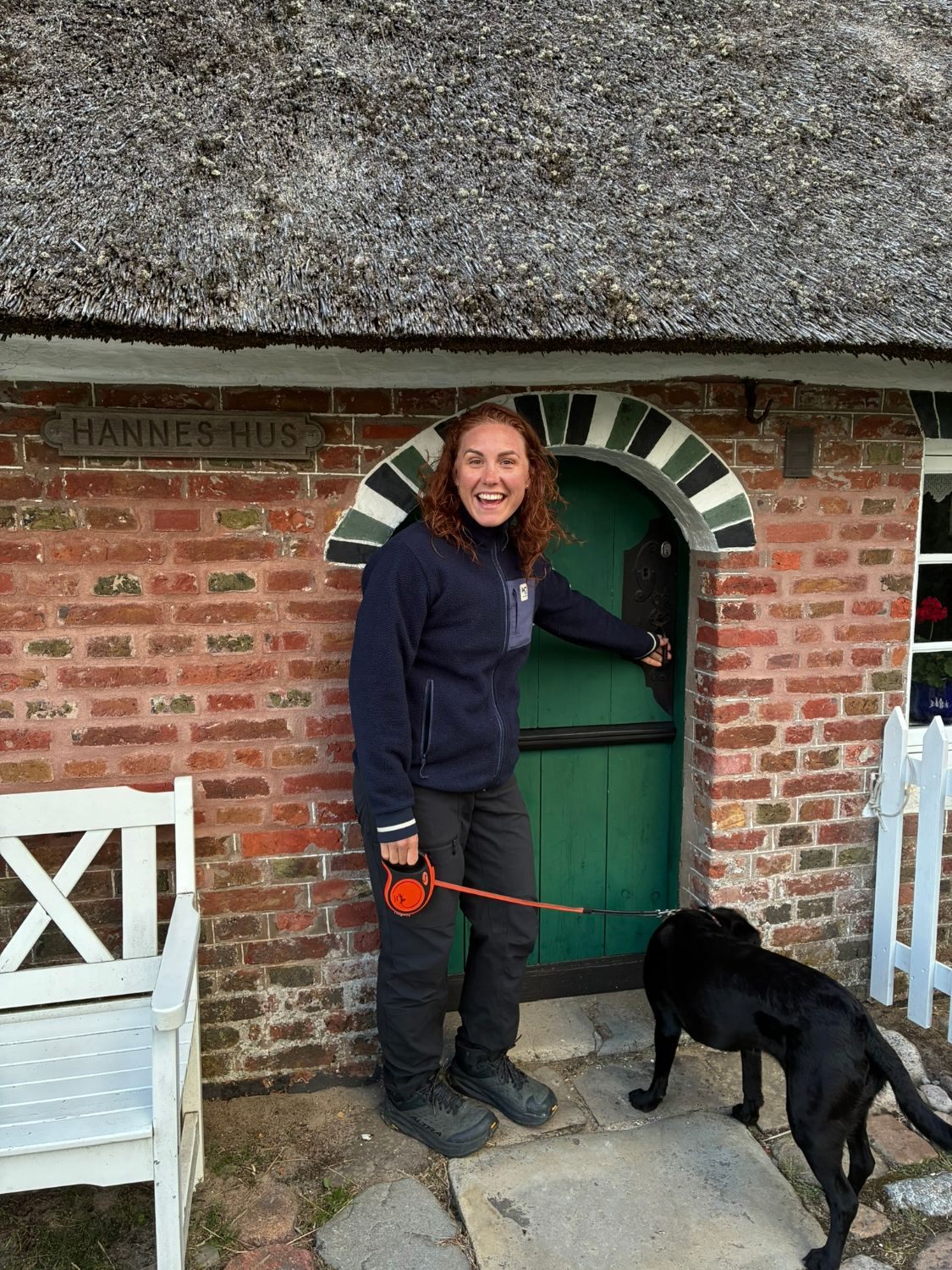 Kristine laughing at the arched green door of Hannes Hus, a thatched red-brick house, with Maggie on the lead