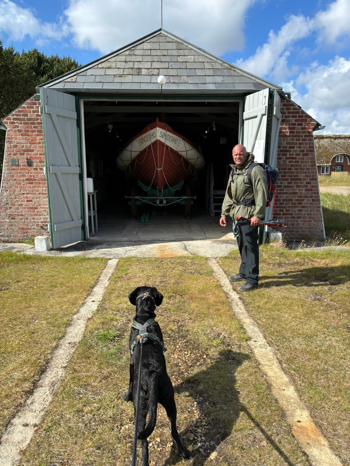 Leif with a backpack outside an old brick boathouse with the red and white Sønderho lifeboat inside