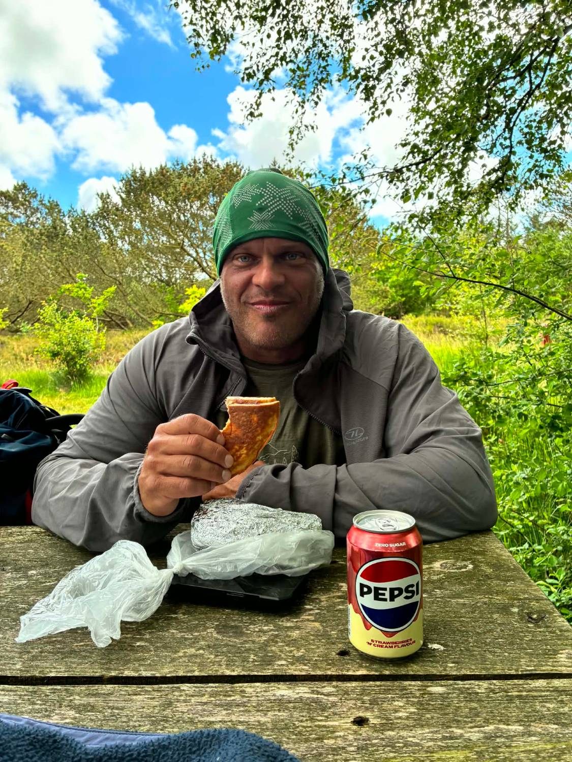 Leif in a green beanie eating a wrap at a wooden picnic table with a can of Pepsi