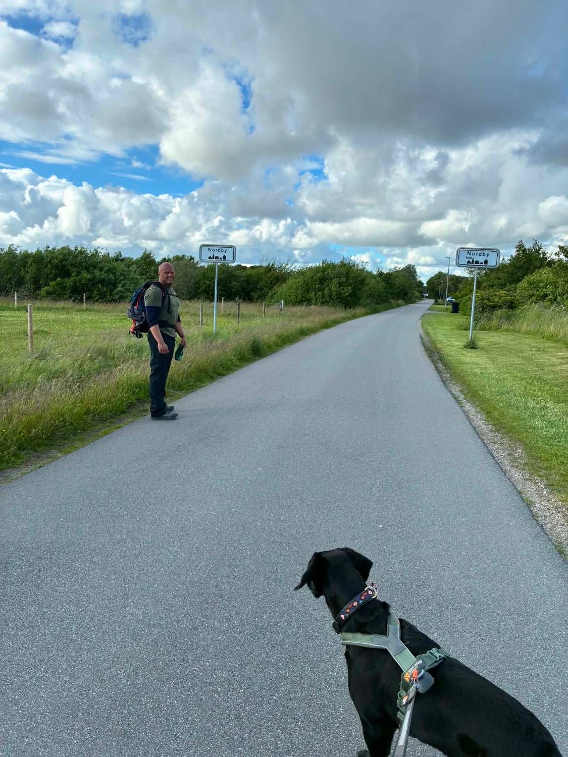 Leif with a backpack by the Nordby town sign on a quiet road, Maggie in the foreground