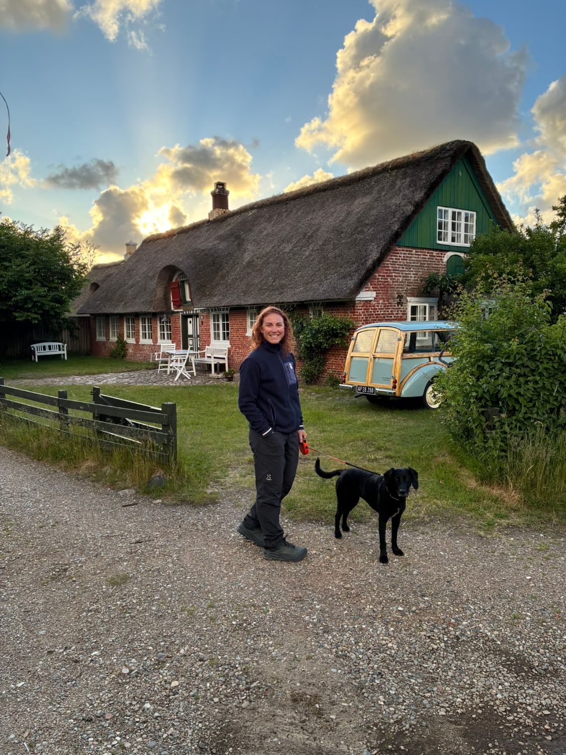 Kristine and Maggie in front of a long thatched red-brick house with a green gable and a vintage wood-framed car, evening sun behind