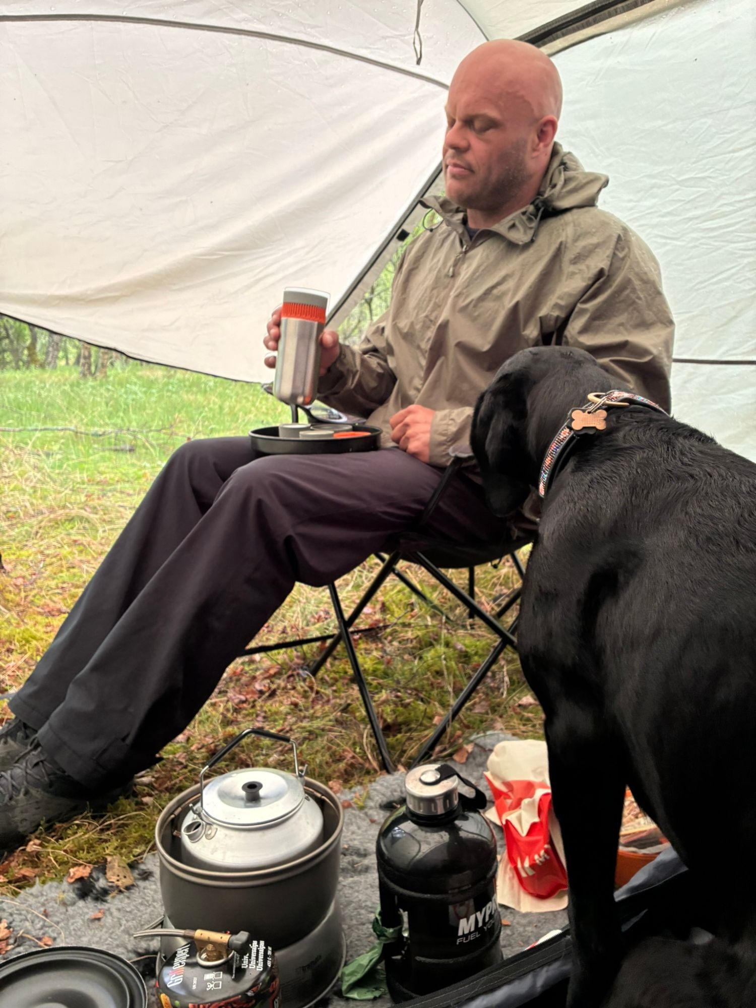 Leif in a rain jacket making coffee in a portable press, sitting in a camping chair under the tent porch with Maggie beside him