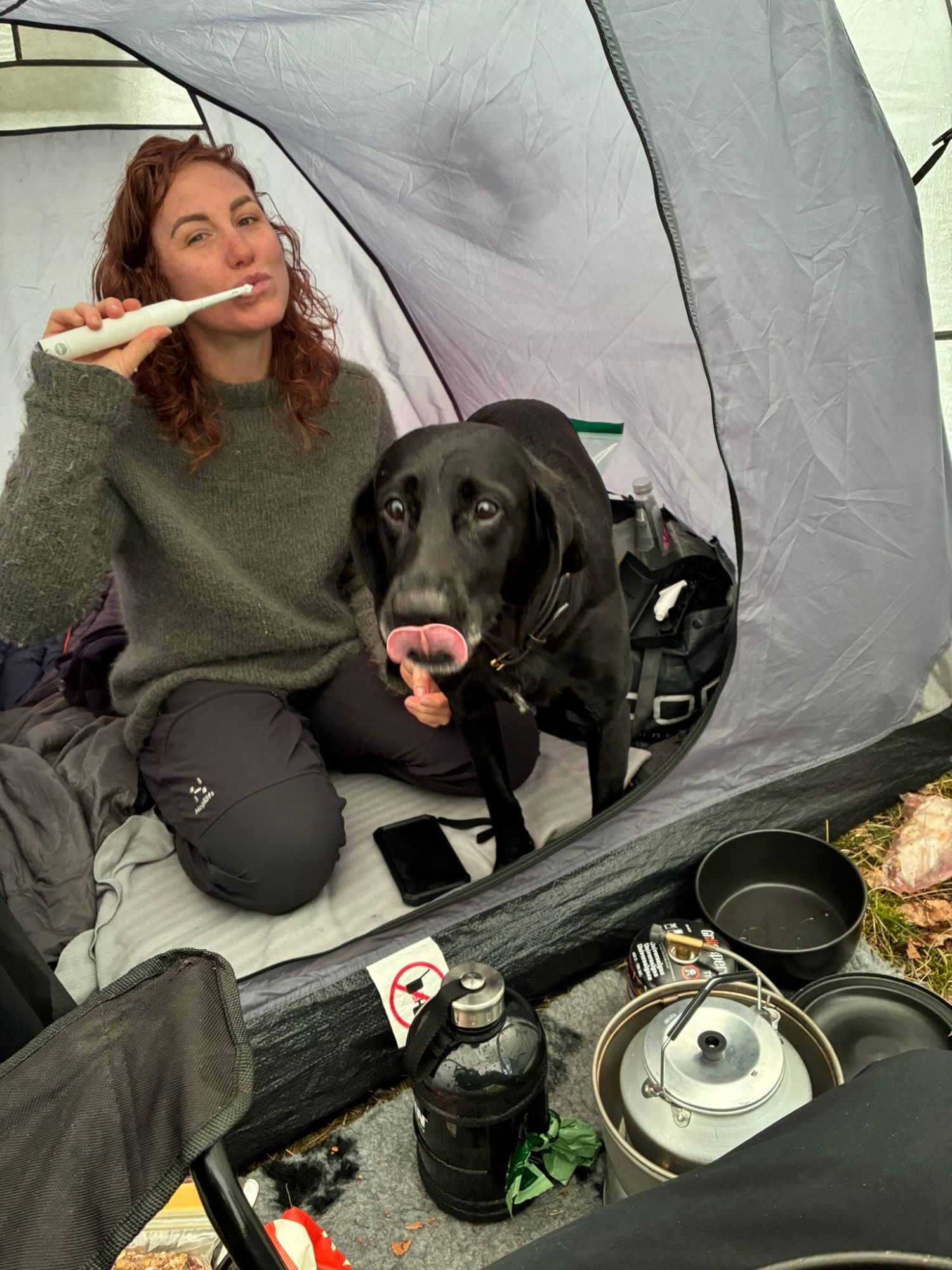 Kristine brushing her teeth inside the tent while Maggie licks her nose next to her