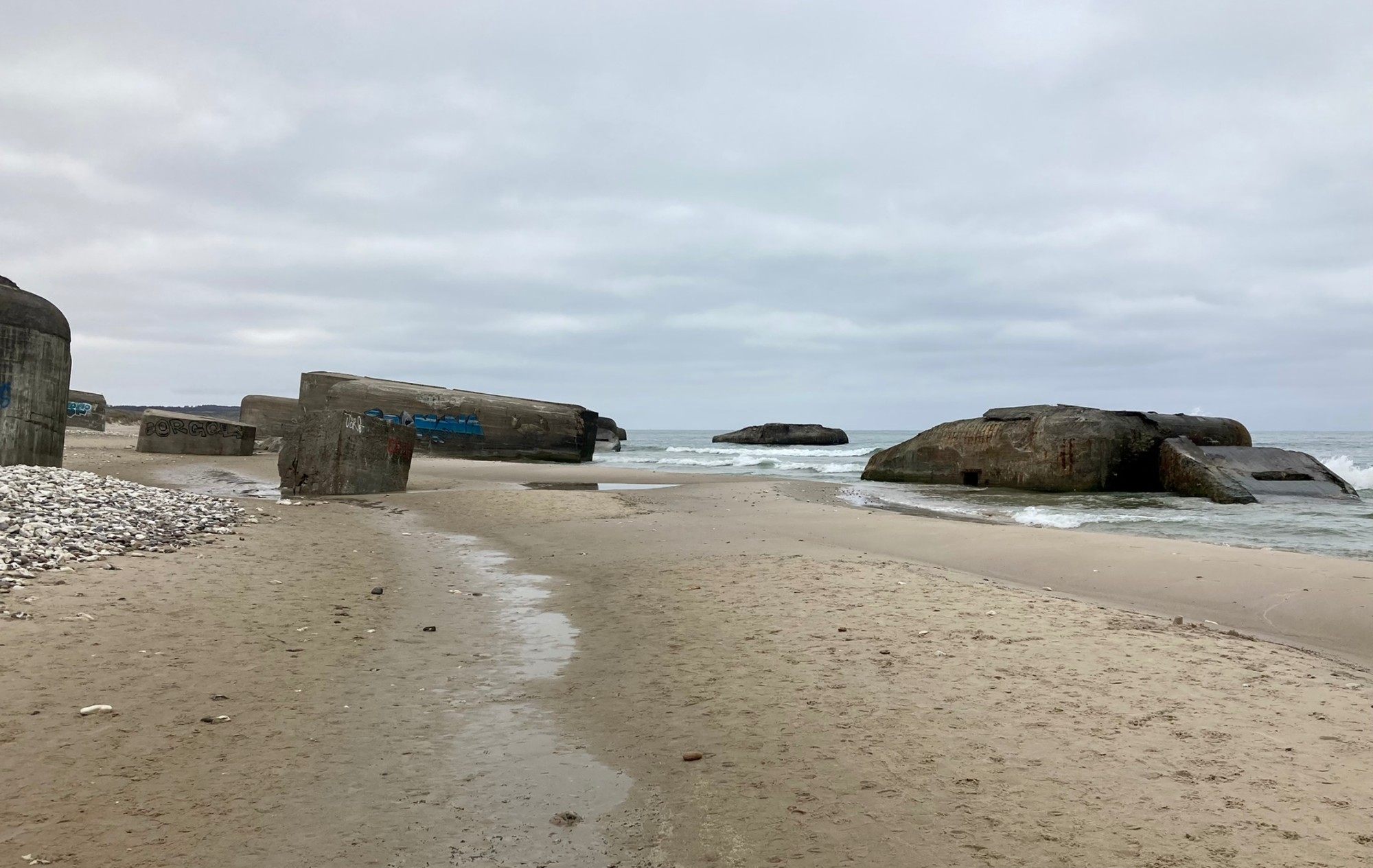 Broken concrete bunkers on the sand and in the surf under a grey sky
