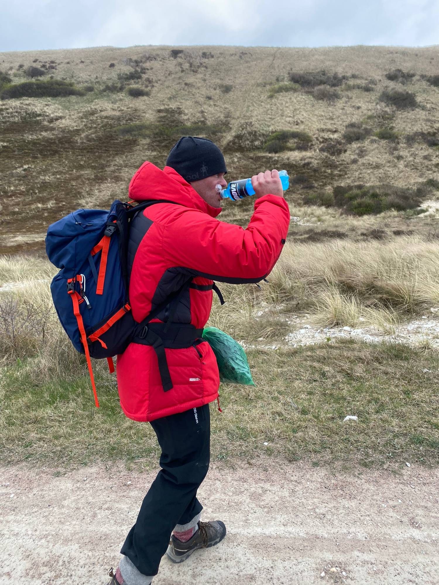 Walking with a backpack and red jacket, drinking from a sports drink bottle on a sandy track below grassy dunes