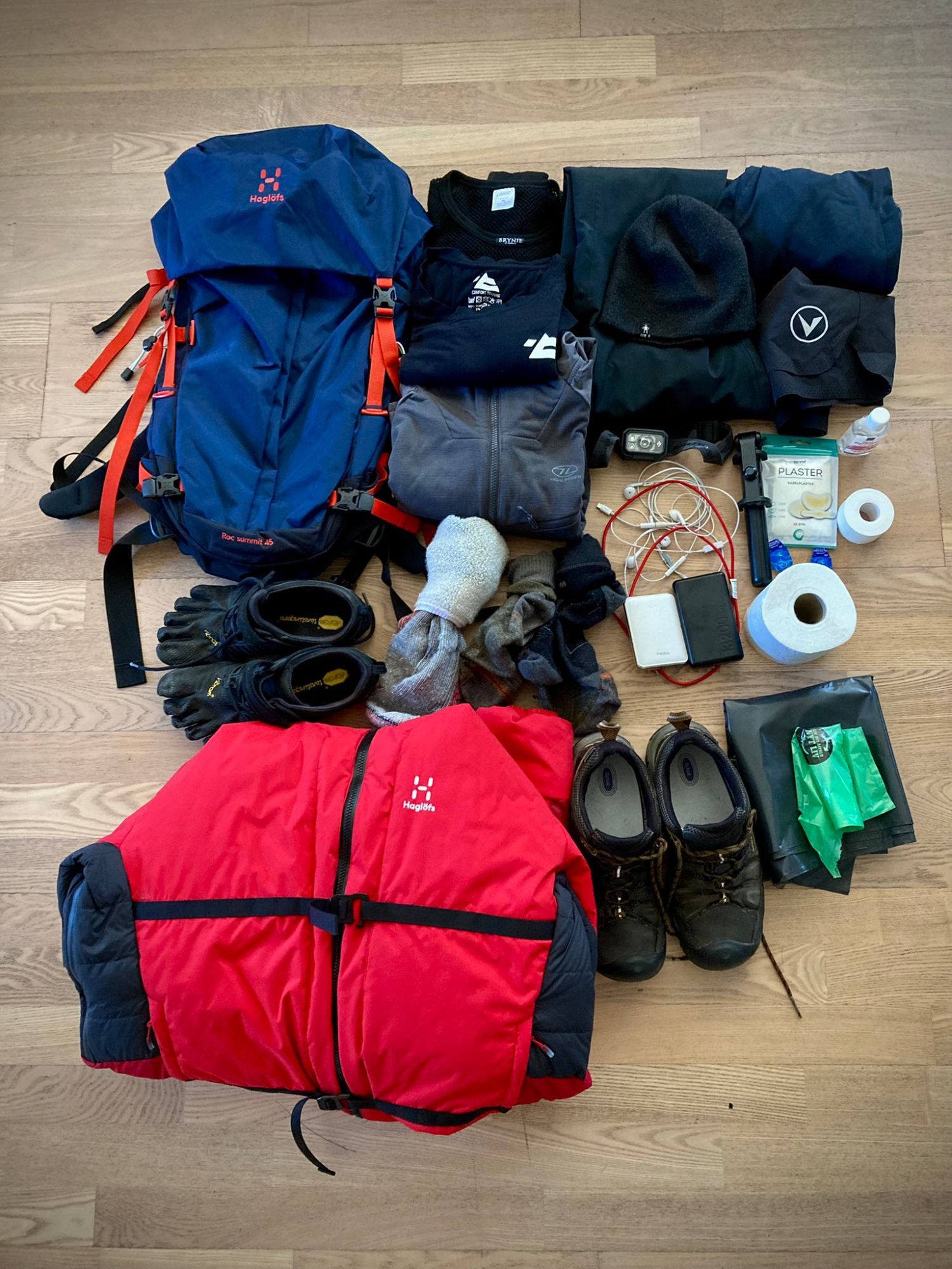 Hiking backpack, red down jacket, clothing, boots, toe shoes, headlamp, power banks and plasters laid out on a wooden floor