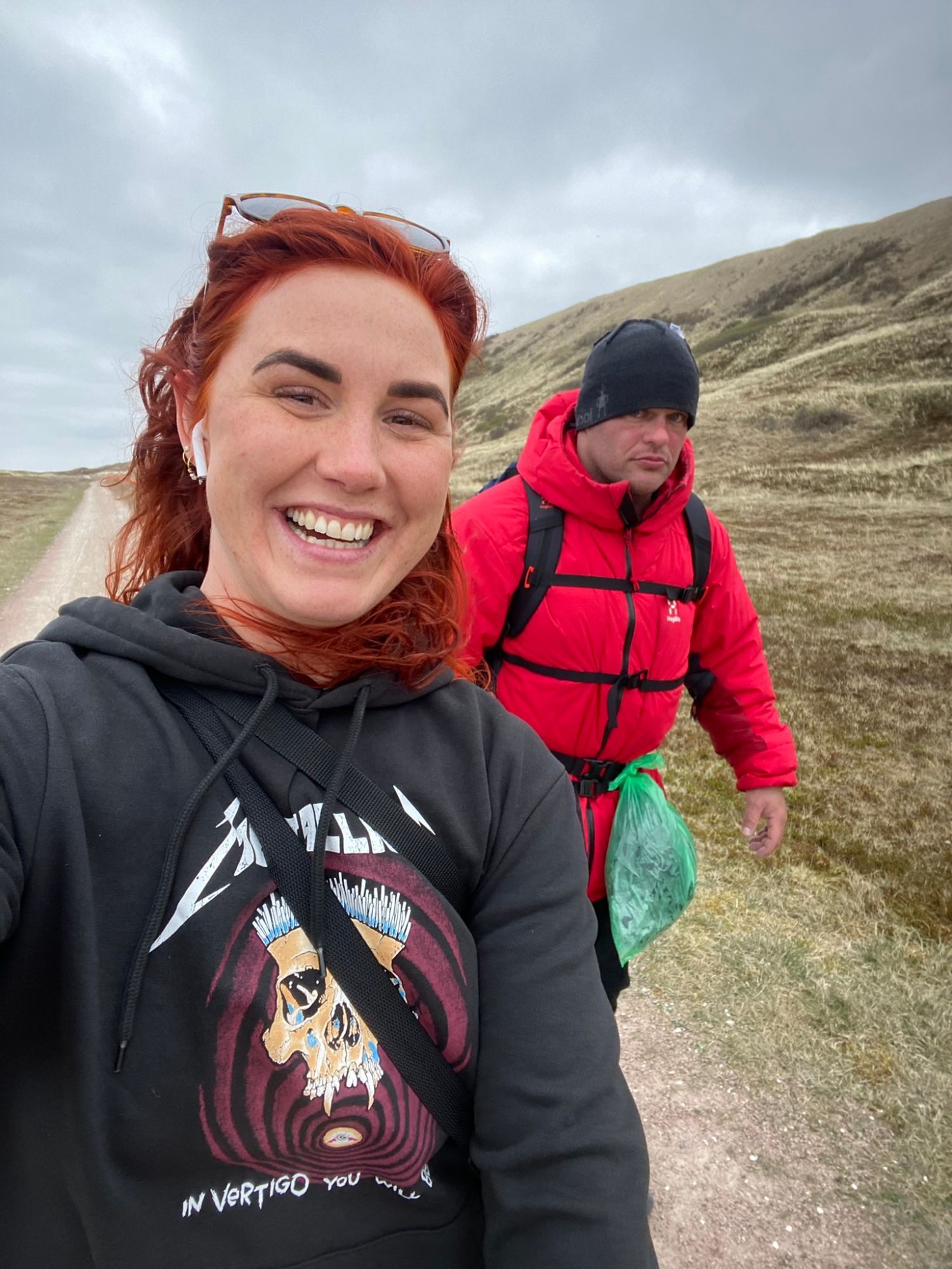 Kristine smiling in a selfie on a sandy track through the dunes, with Leif behind in a red jacket looking exhausted