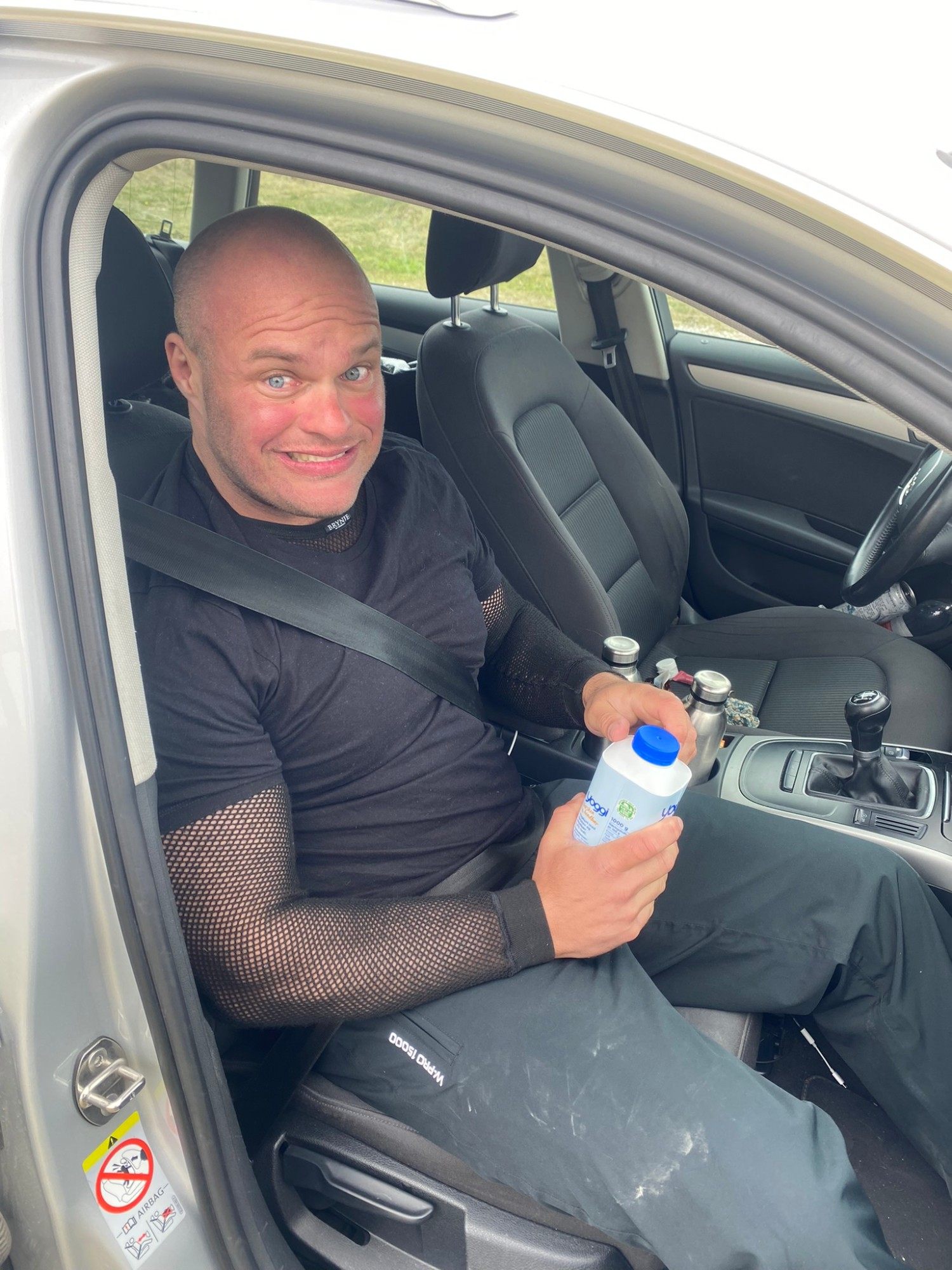 Grinning in the passenger seat of a car holding a carton of drinking yoghurt
