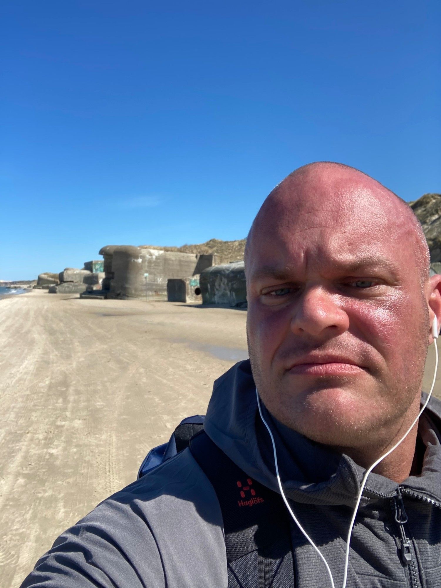 Sunburnt selfie with white earphones in front of a row of bunkers on the beach