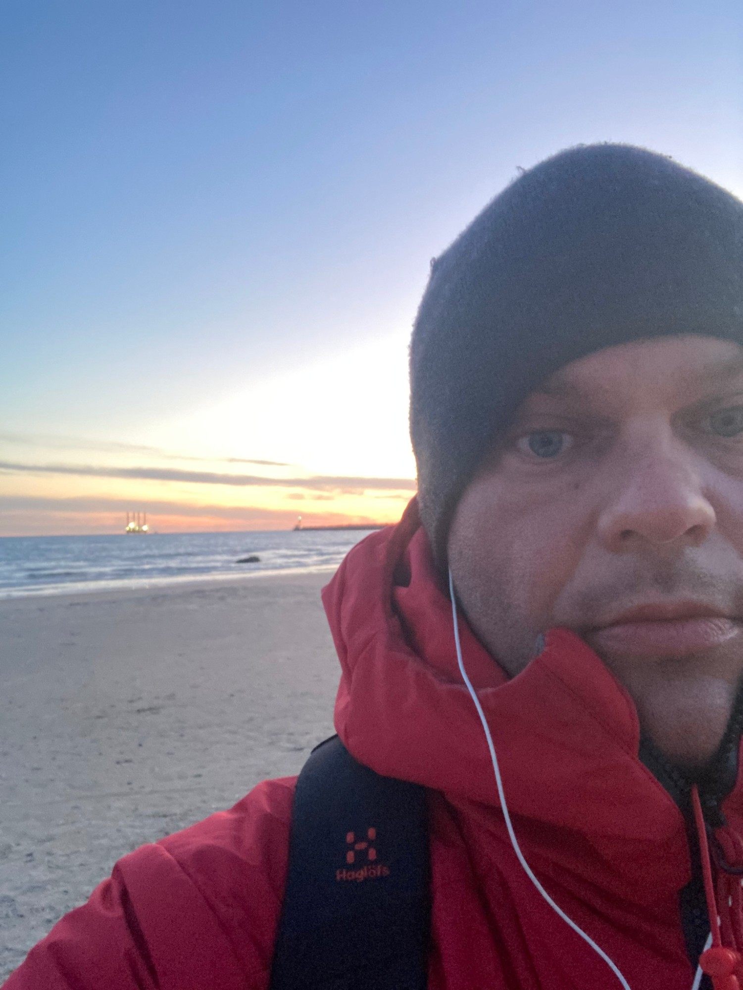 Selfie in a red jacket and beanie on the beach at Hirtshals at dawn, with the harbour lights behind