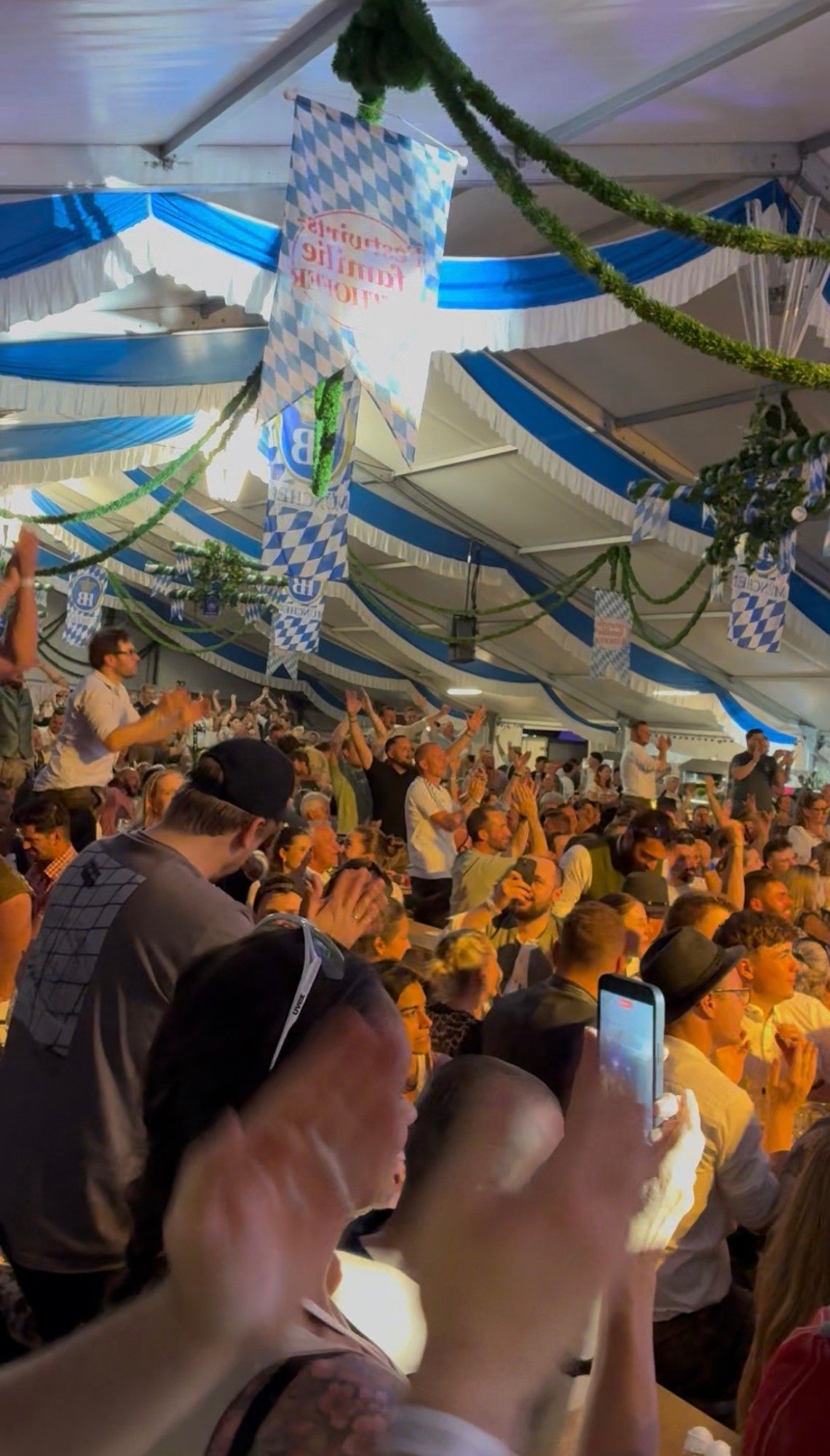 The crowd in the Bavarian festival tent on its feet, clapping and cheering under blue and white decorations