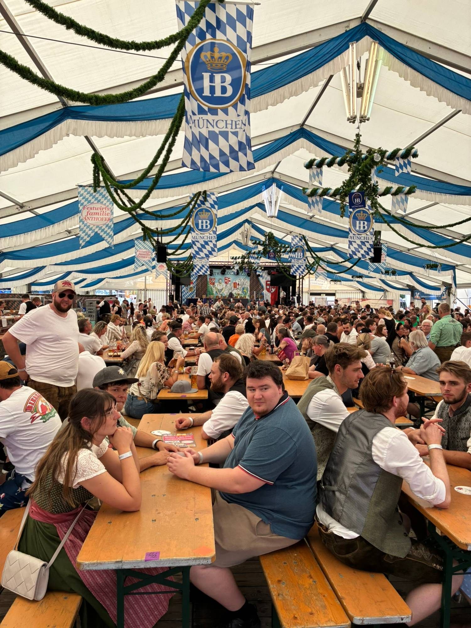 A packed Bavarian festival tent with blue and white flags, long tables full of people and the stage at the far end
