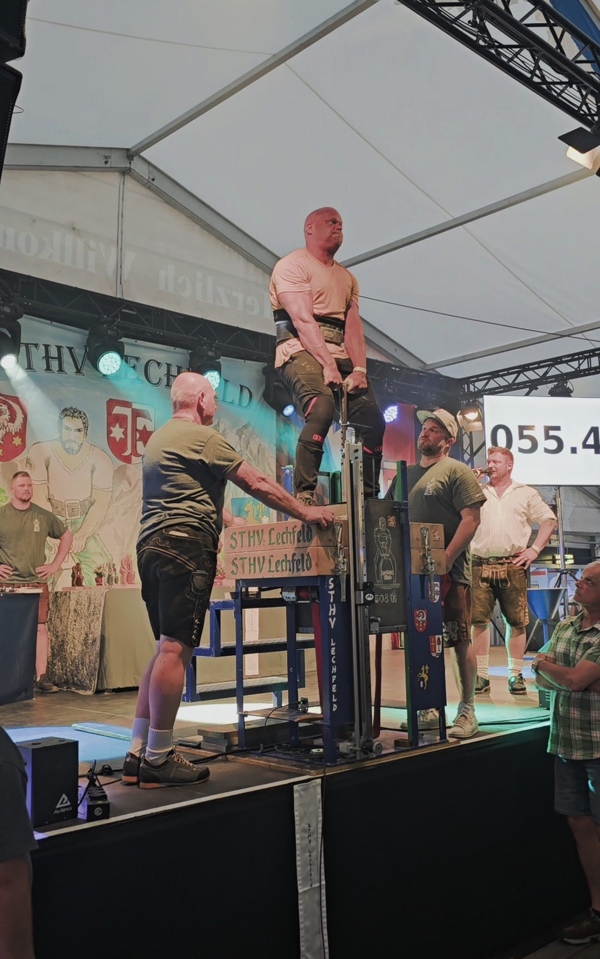 Standing upright on the platform holding the 254 kg stone, with the screen behind showing 055.4