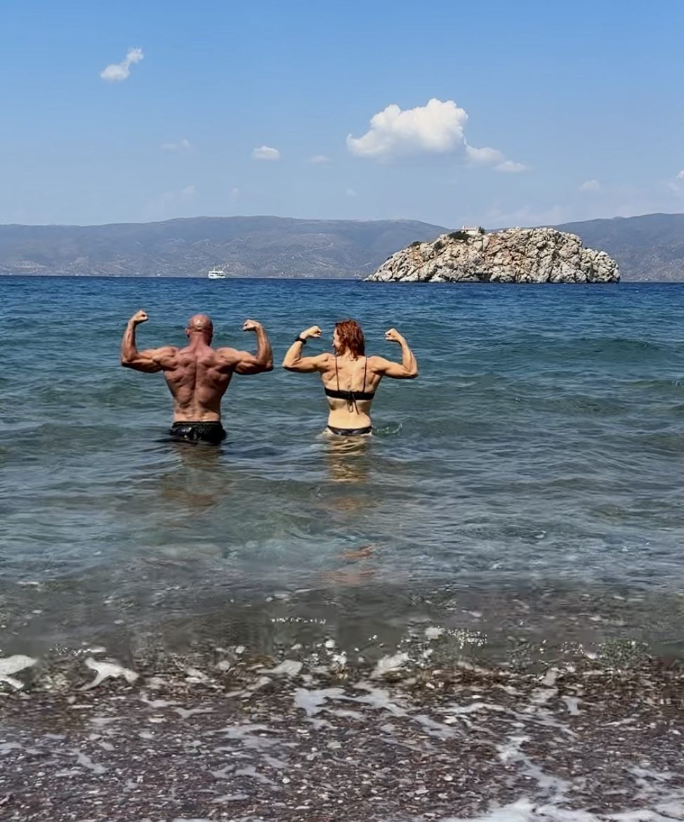 The two of us flexing in the clear sea off Hydra, with a rocky islet and the mainland behind