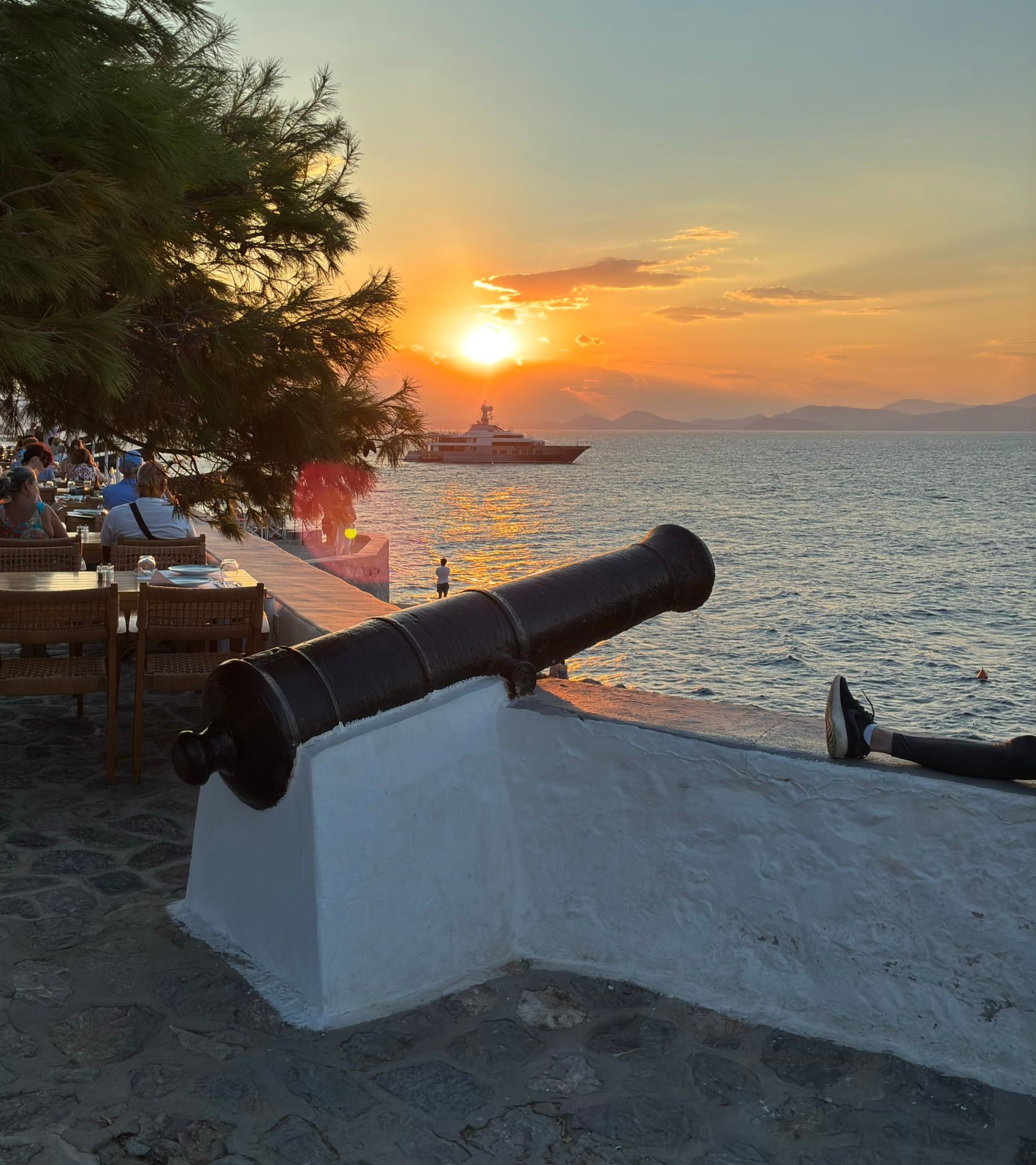 An old cannon on the harbour wall of Hydra pointing out to sea at sunset, with a yacht in the bay