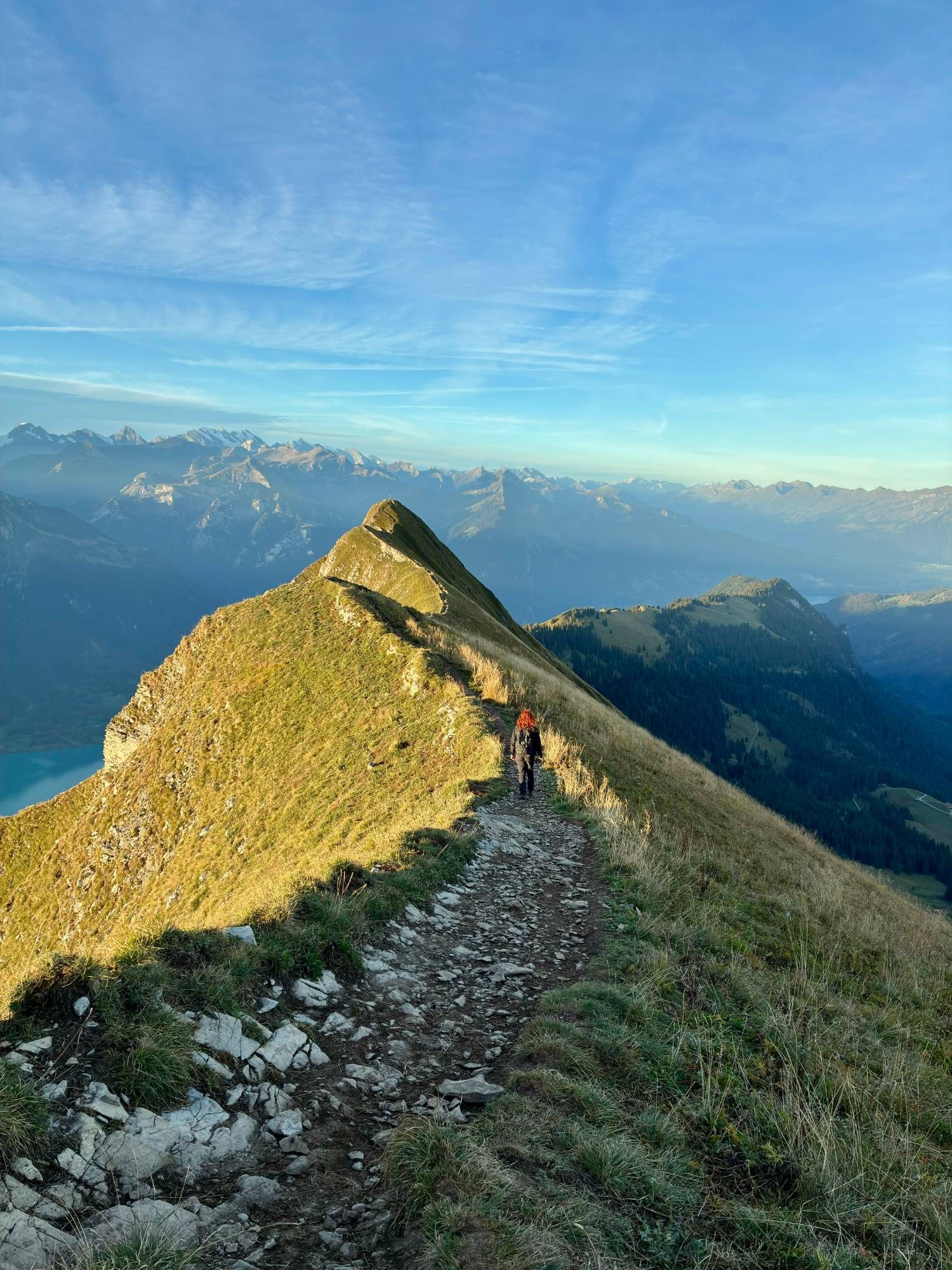 Kristine walking along the narrow ridge path in the morning sun with the Alps behind