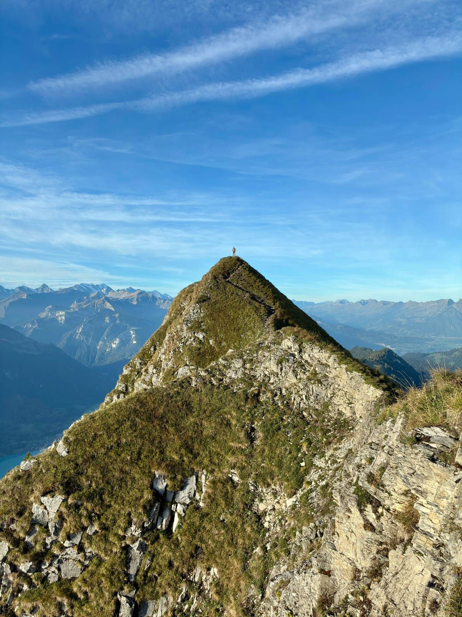 A tiny figure standing on top of a pointed grassy peak along the ridge, with the Alps behind