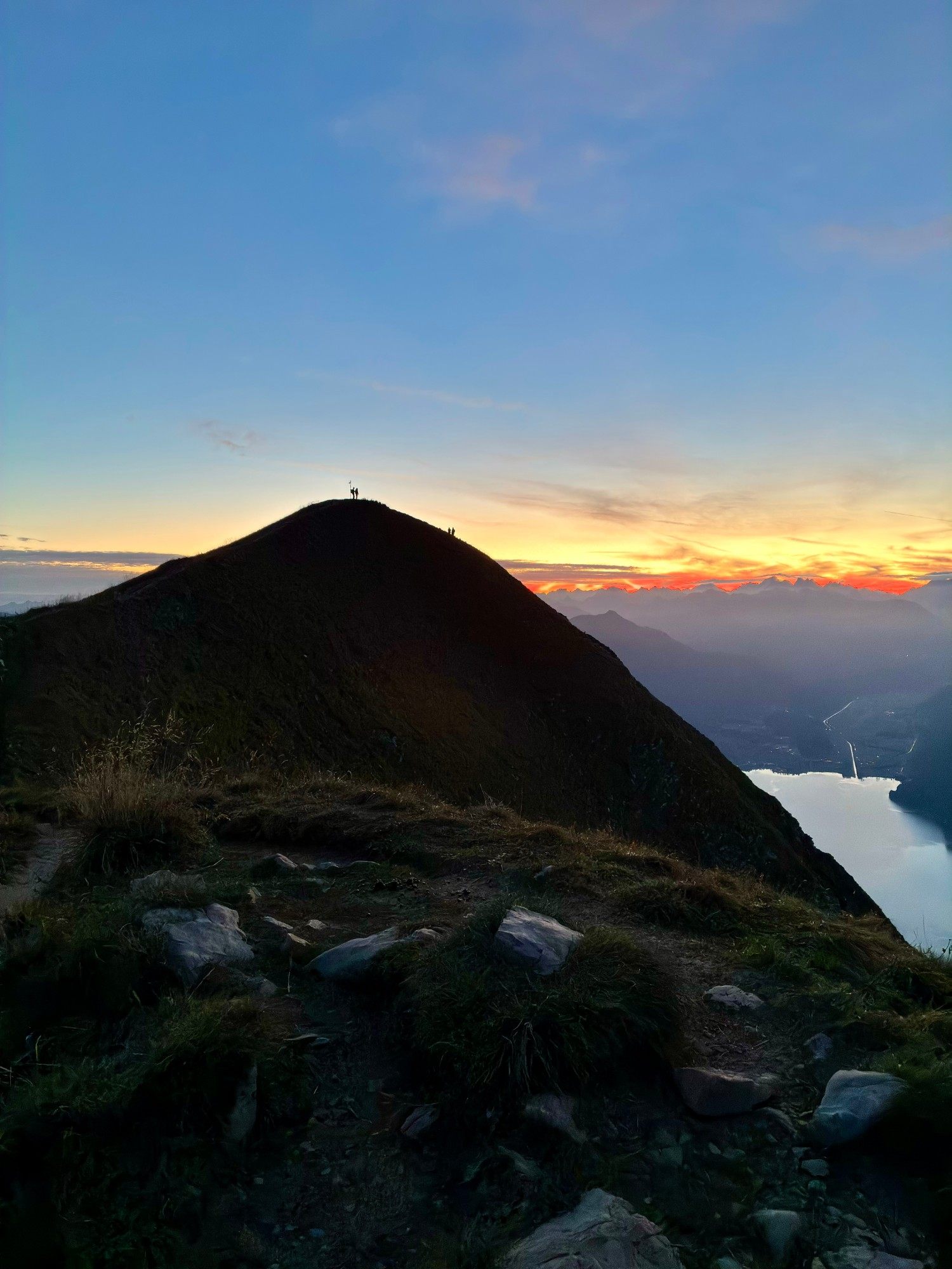 The summit of the Augstmatthorn in silhouette at dawn, with a glowing orange sky over the Alps and Lake Brienz below