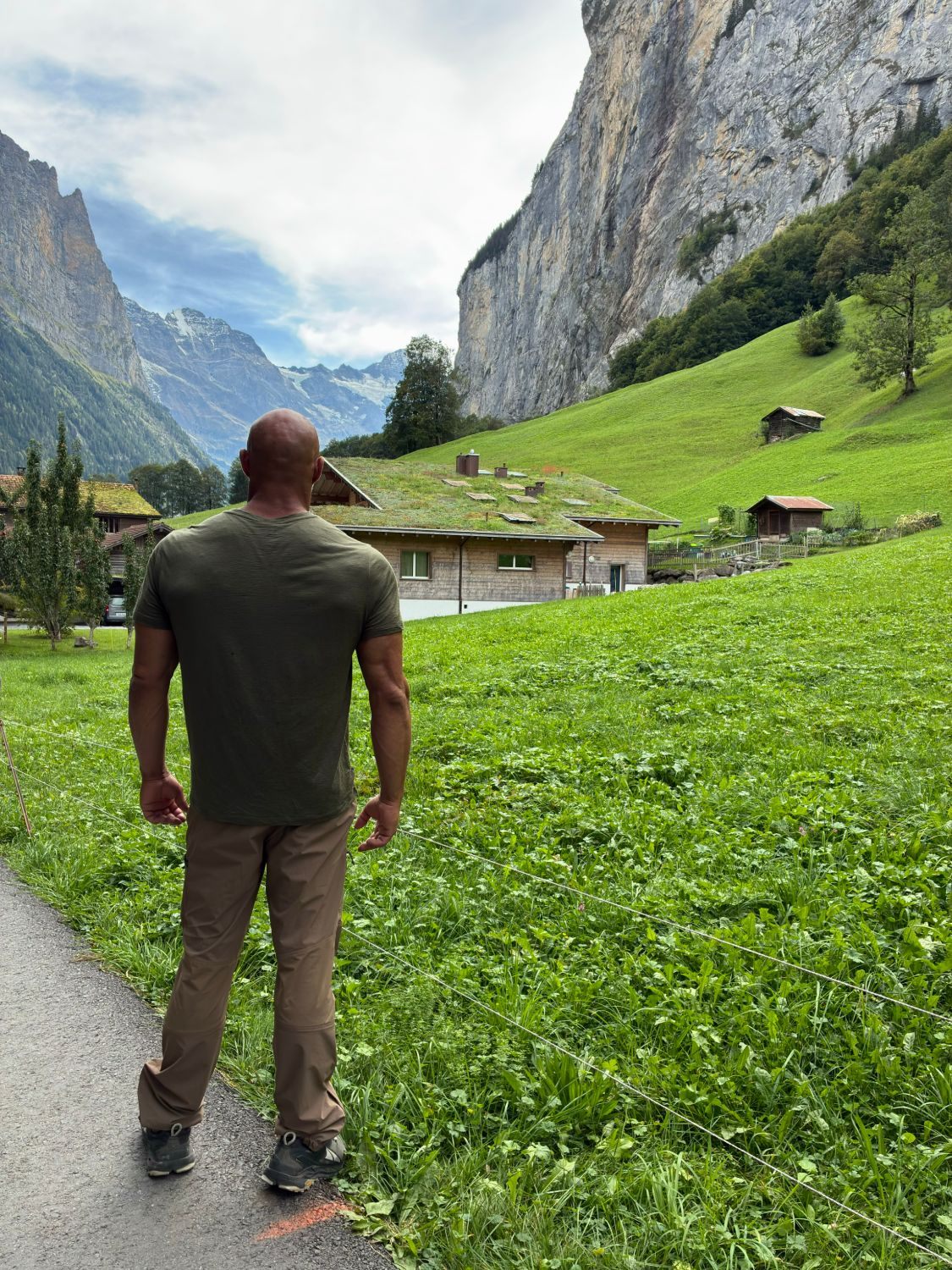 Standing on the path beside a green meadow and a house with a grass roof, under a huge cliff