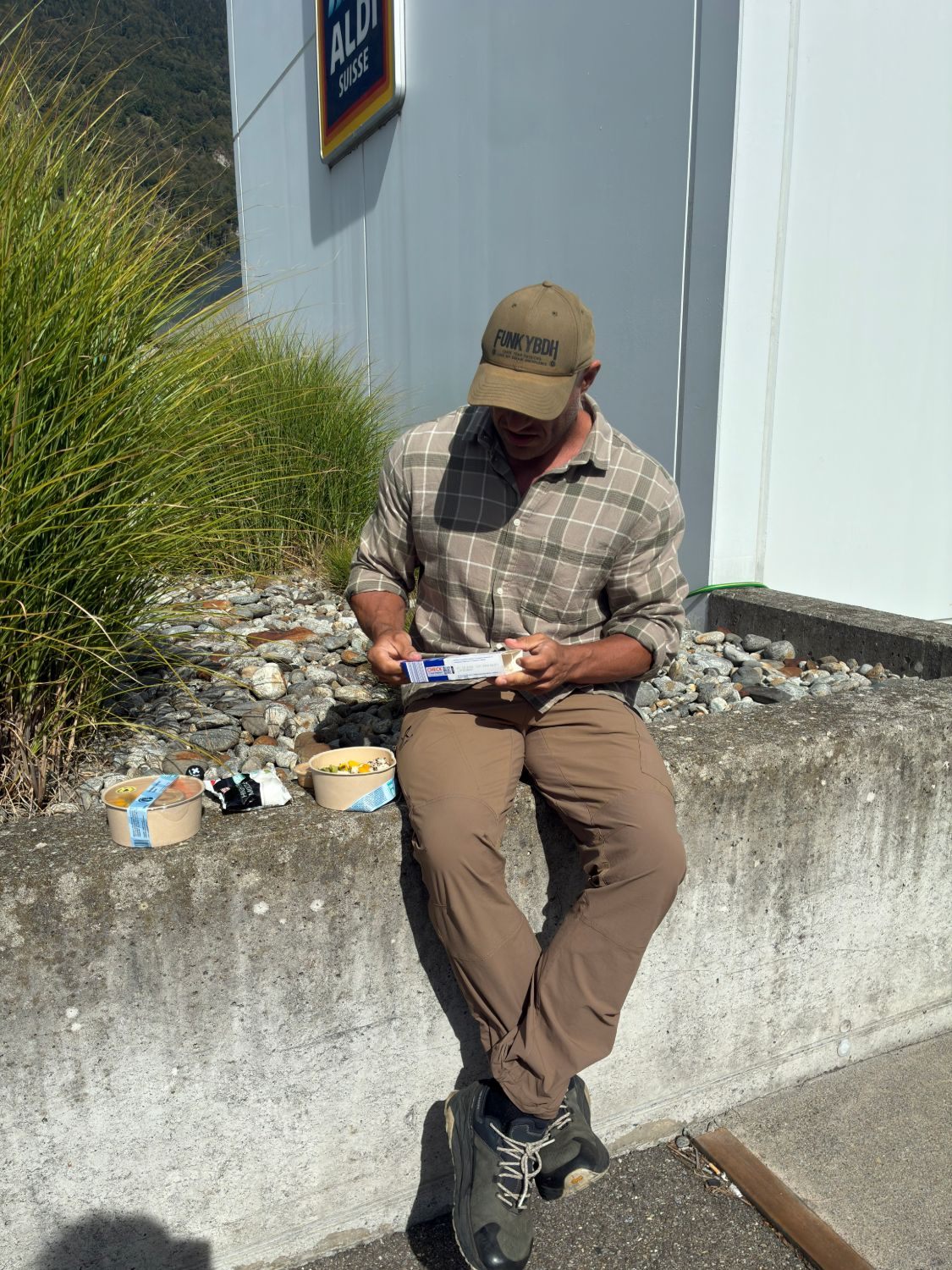 Leif sitting on a low concrete wall outside a supermarket in Interlaken with lunch boxes beside him