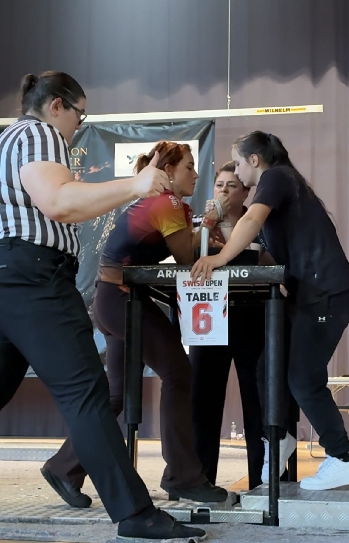 Kristine gripped up on the right arm at table 6 against the athlete who later won the right-arm final, with two referees setting the grip