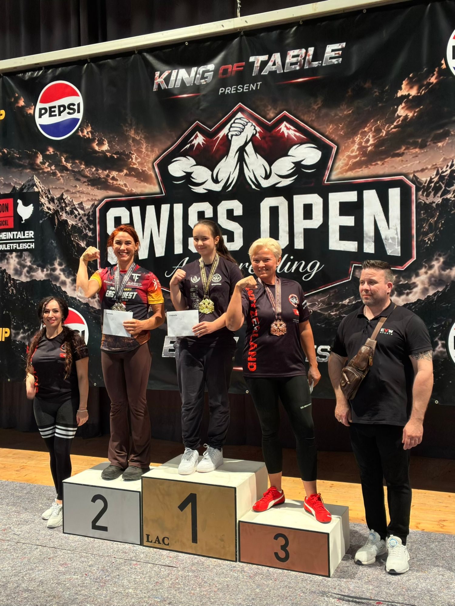 Kristine flexing on the second step of the Swiss Open podium with a silver medal, next to the winner and the bronze medallist