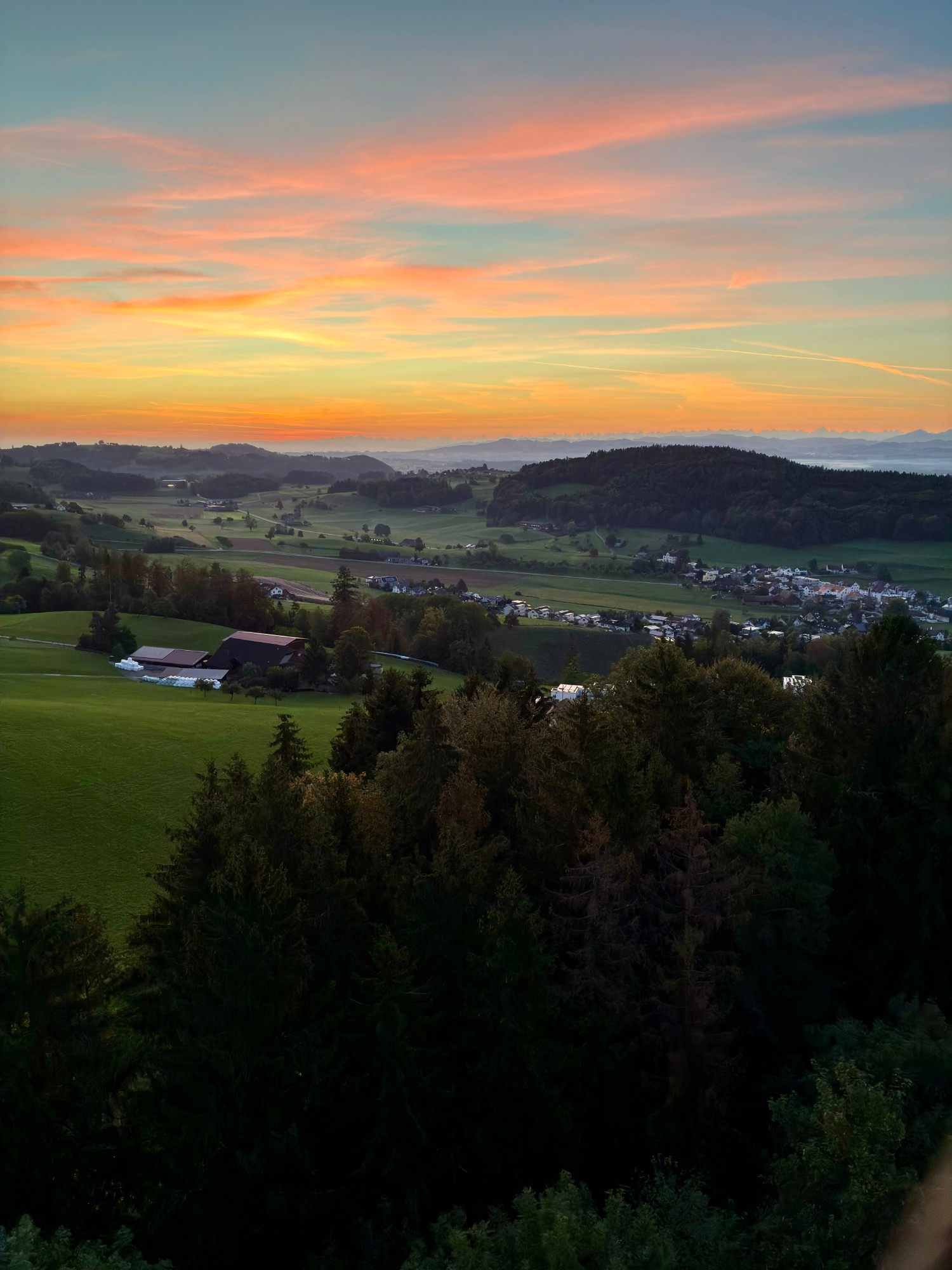 Orange sunrise sky over green fields, woods and a village, with a faint line of mountains on the horizon