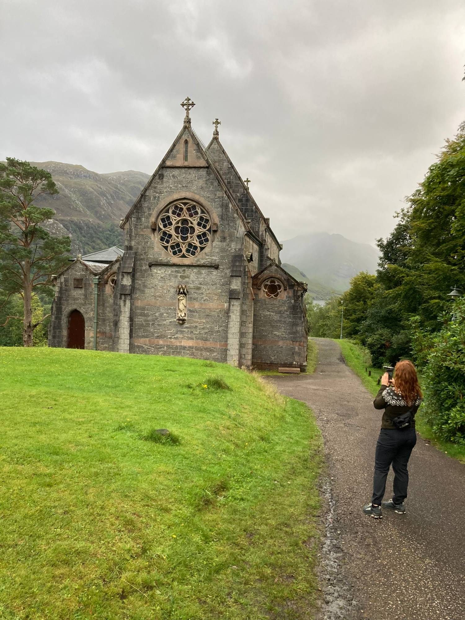Kristine photographing the grey stone church of St Mary and St Finnan in Glenfinnan, with misty mountains behind