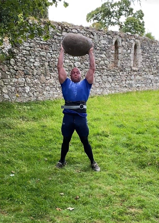 Leif pressing the Barevan Stone overhead with straight arms in front of the old church wall