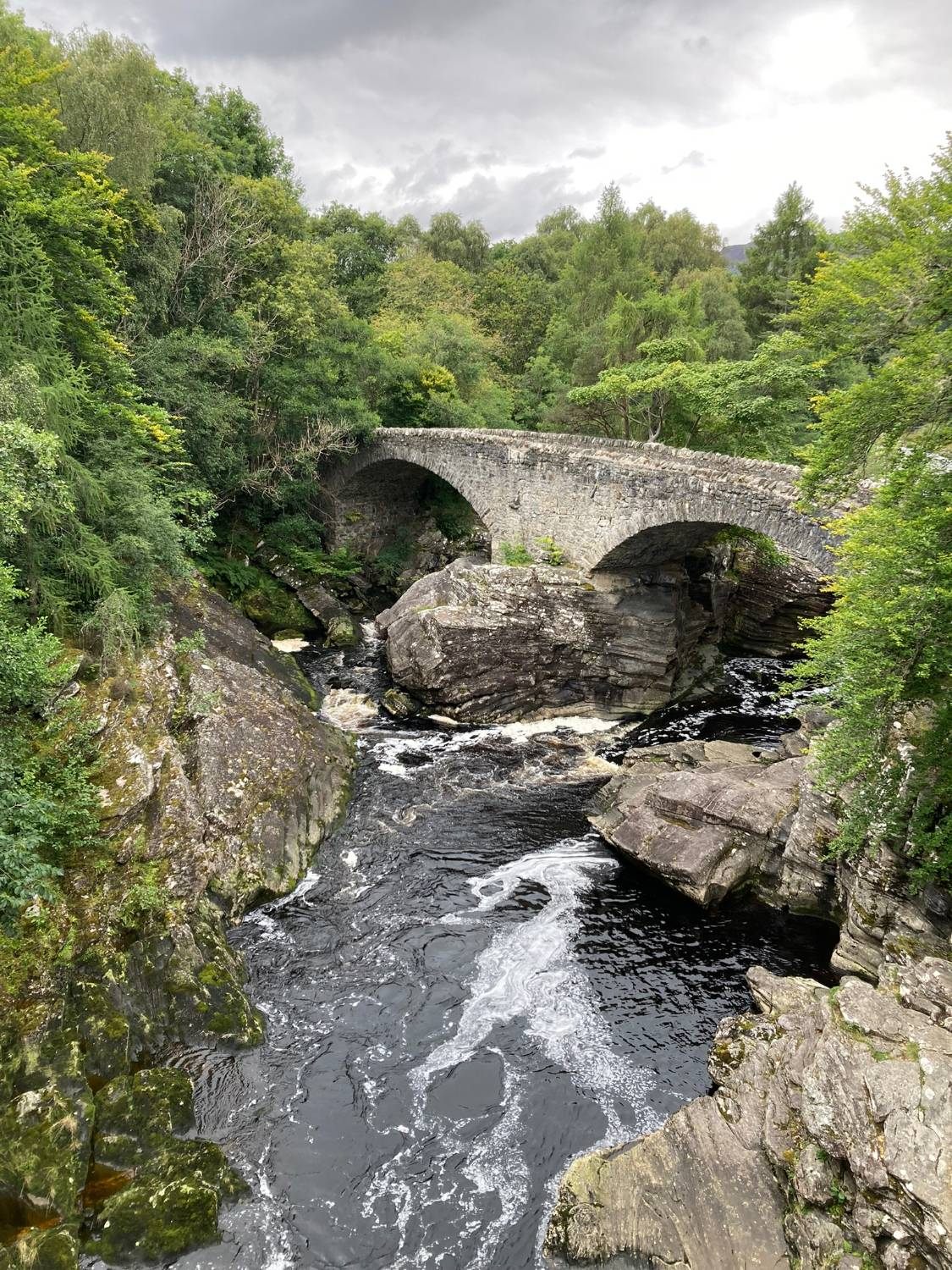 The old two-arched stone bridge at Invermoriston over dark water and rocks, surrounded by trees