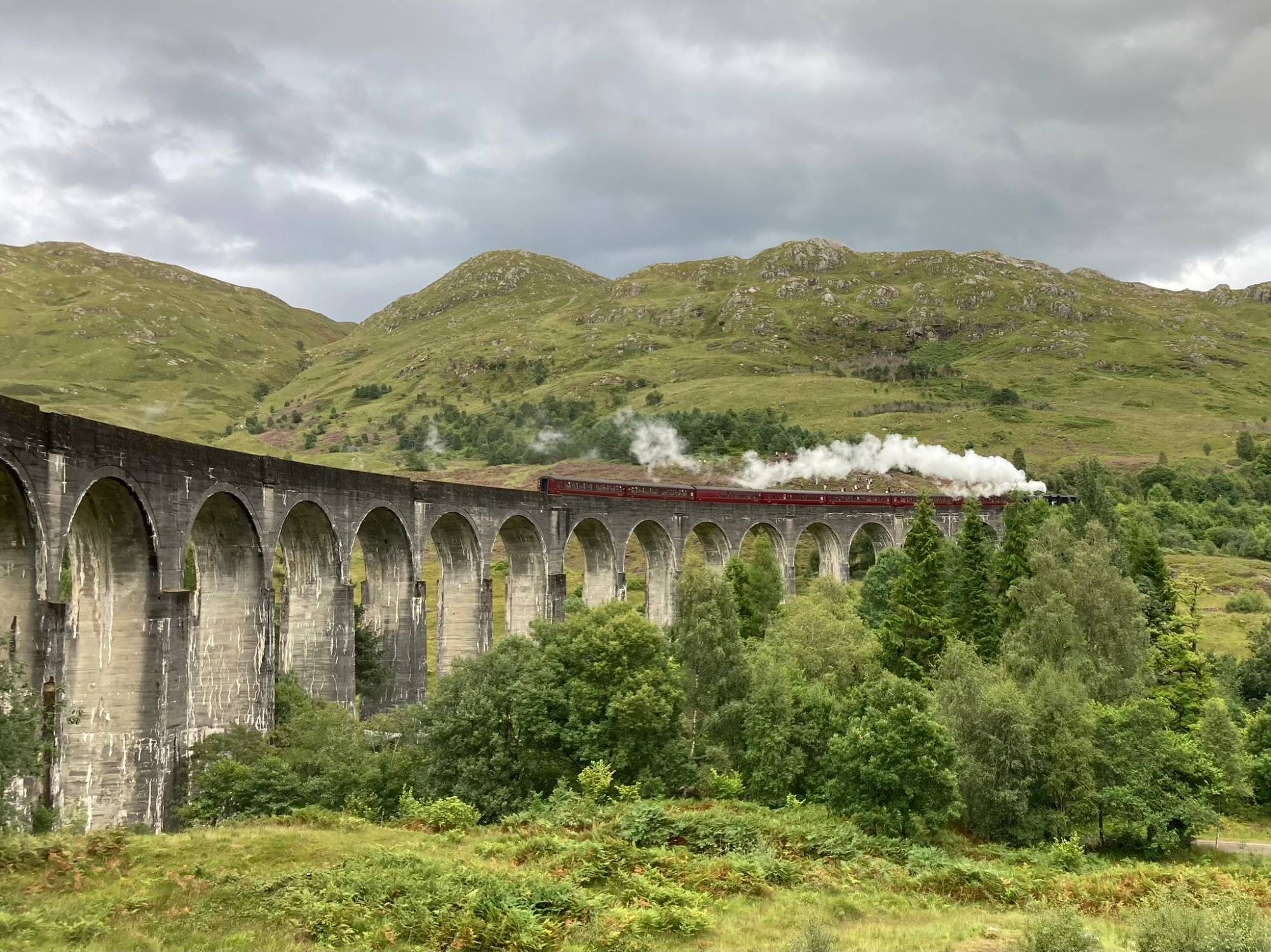 The Jacobite steam train with maroon carriages crossing Glenfinnan Viaduct, white steam trailing above