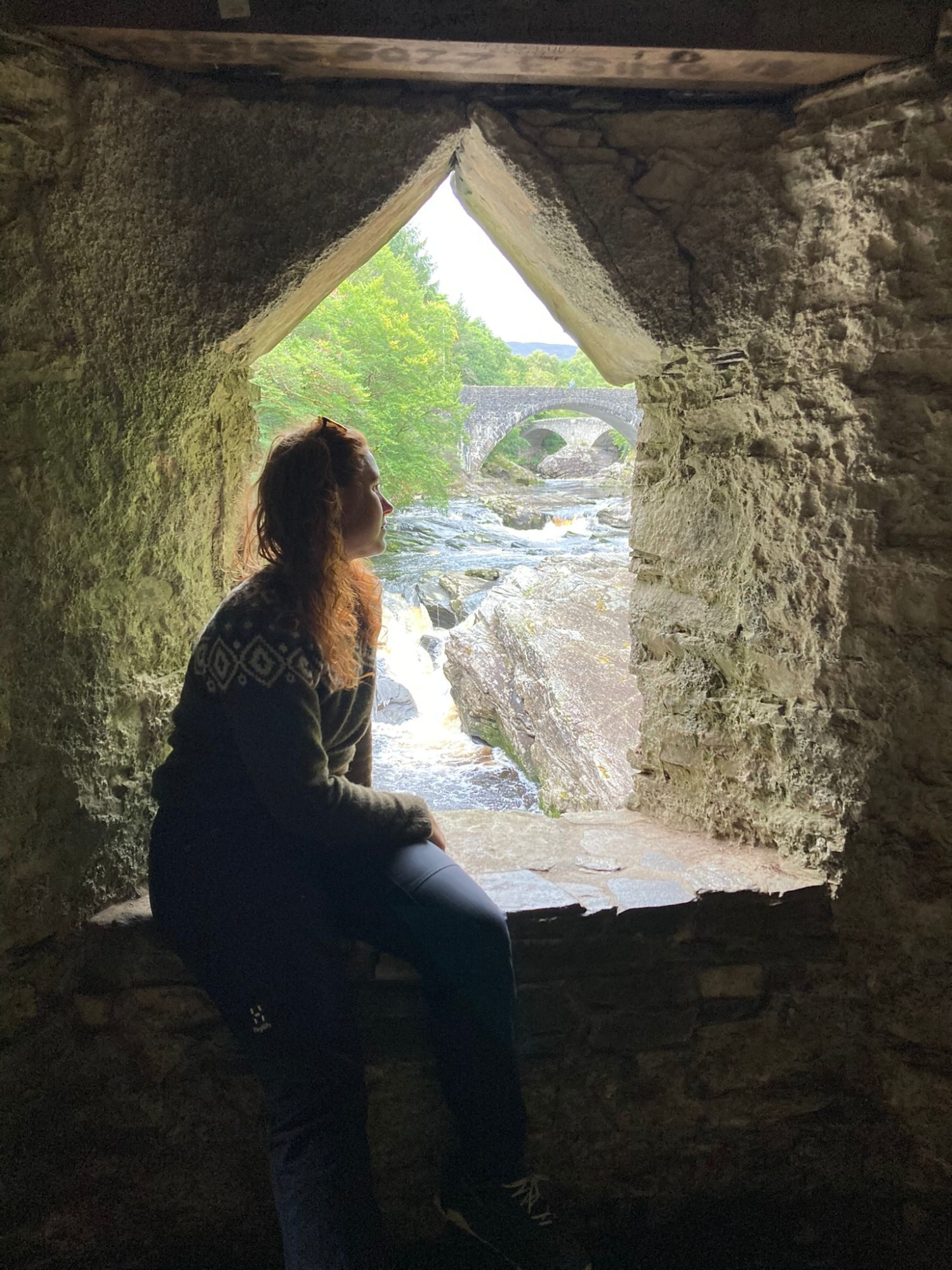 Kristine silhouetted in a pointed stone window looking out at the falls and the bridge