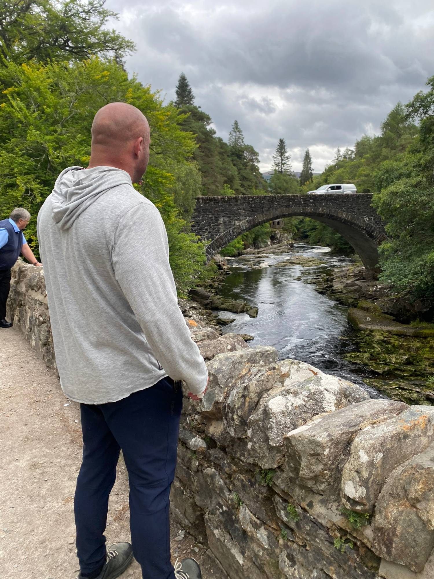 Leif in a grey hoodie looking over the stone parapet at the newer road bridge over the River Moriston