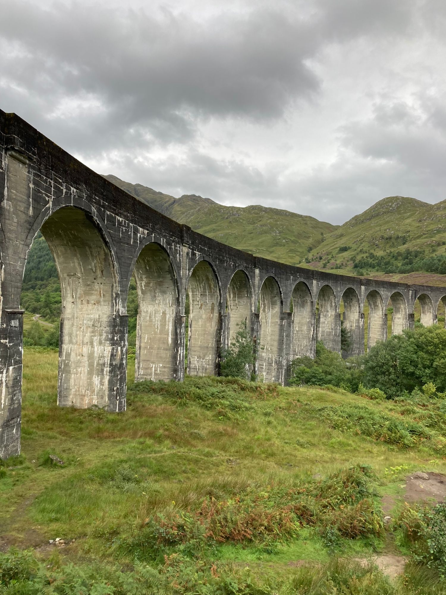 The curving concrete arches of Glenfinnan Viaduct with green Highland hills behind under grey cloud