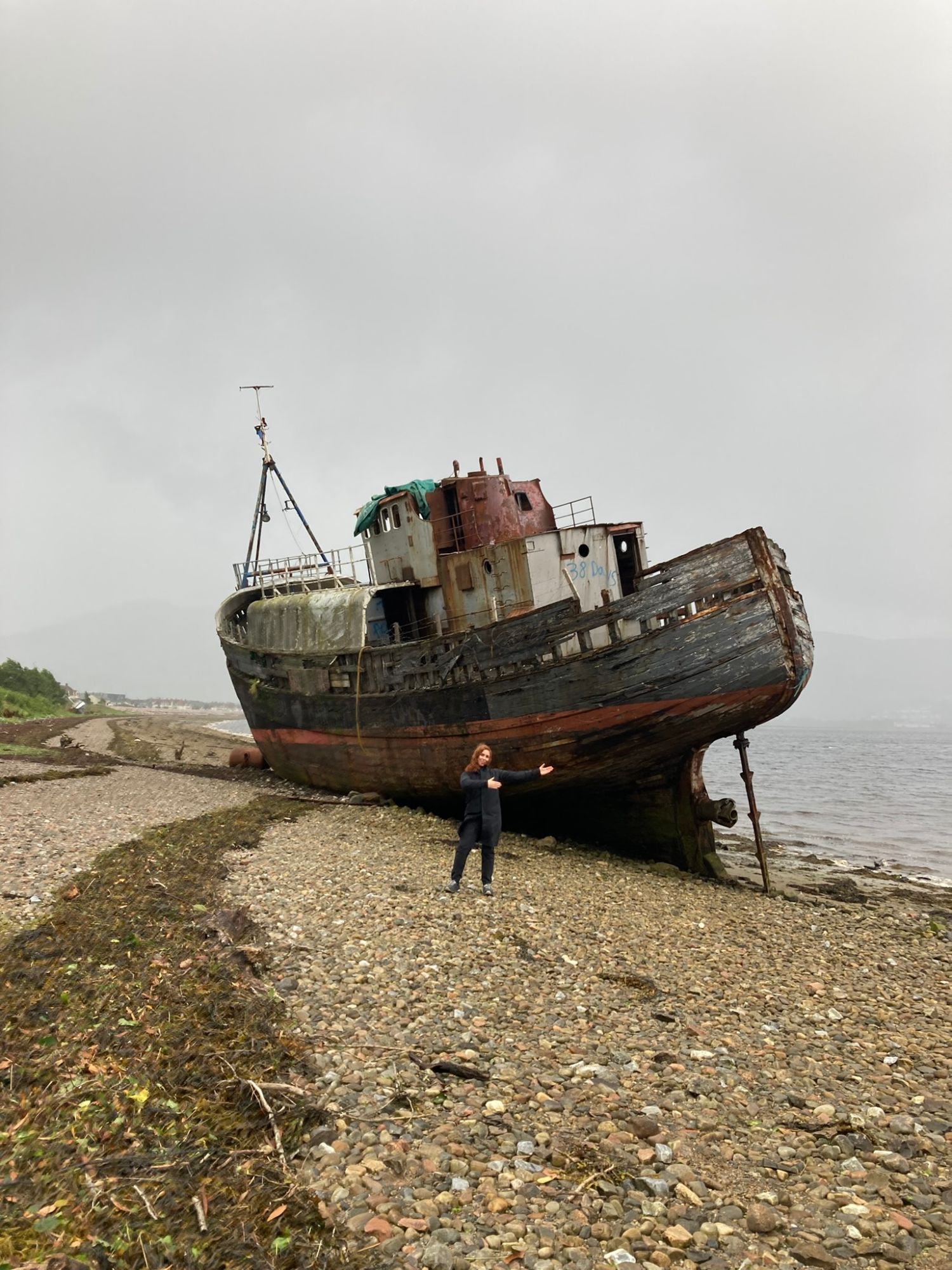 Kristine with arms out in front of the stranded fishing boat known as the Corpach Wreck on the stony shore of Loch Linnhe