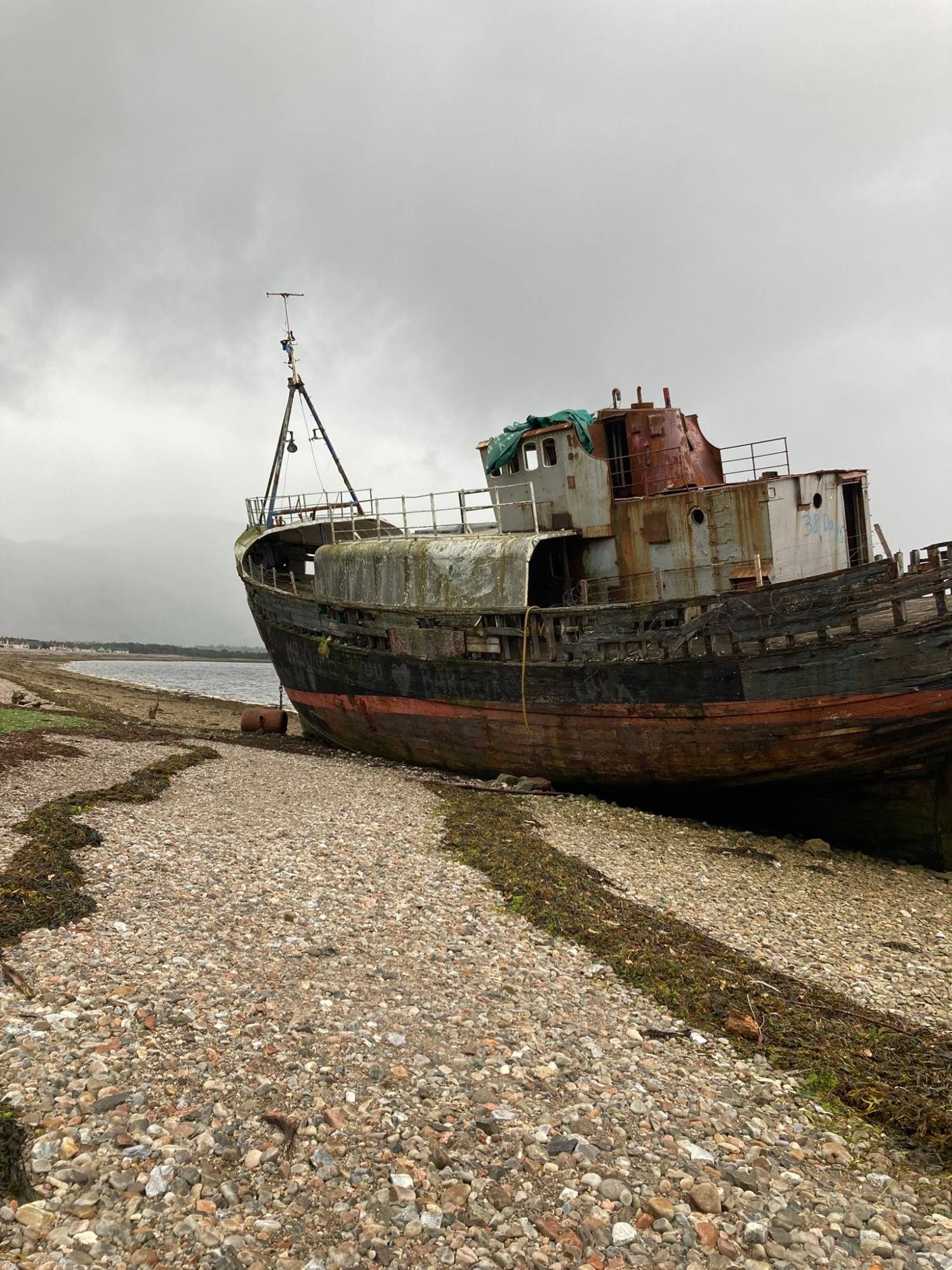 The weathered wooden hull and rusting wheelhouse of the Corpach Wreck on a pebble beach