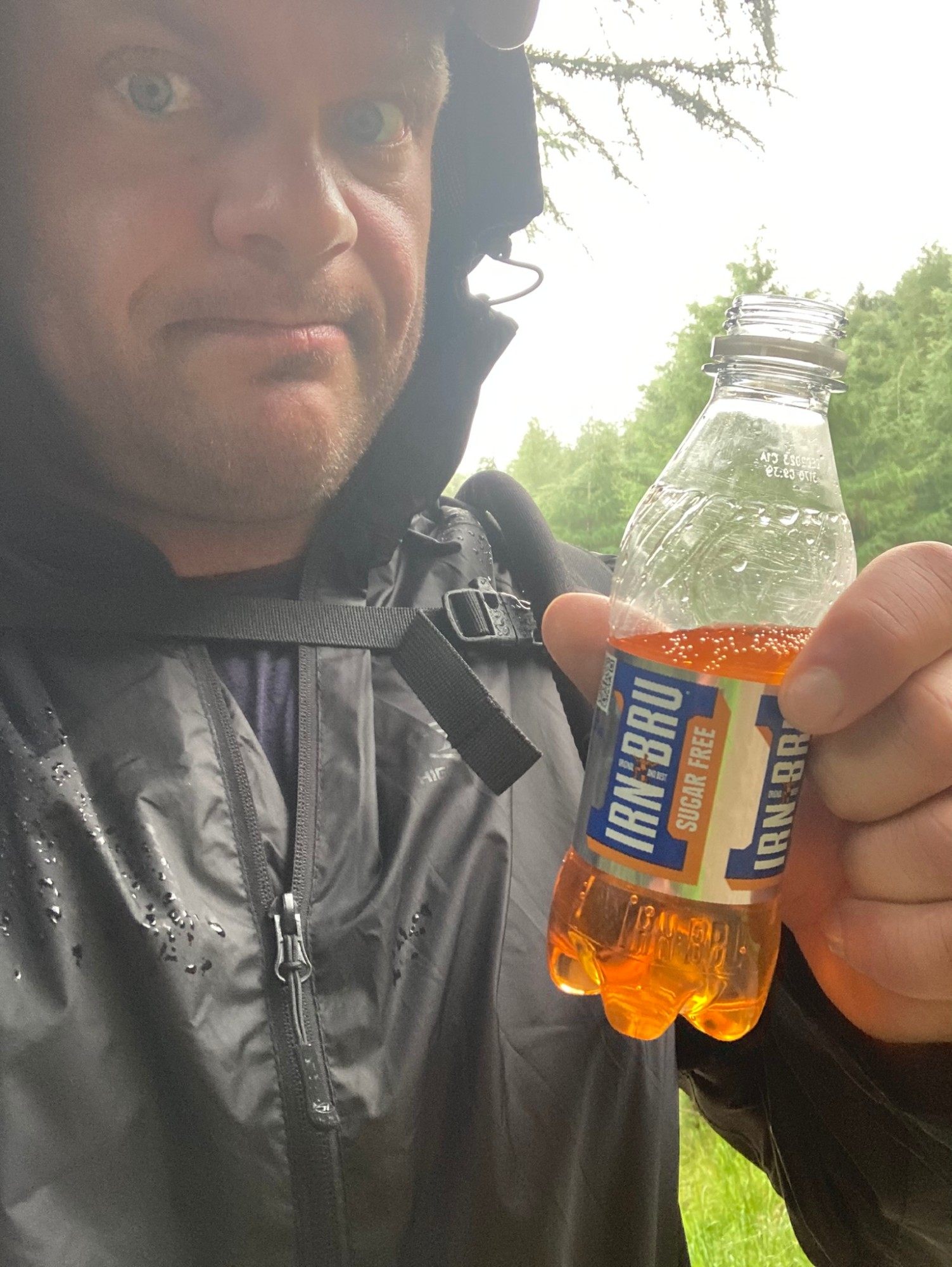 Leif in a wet hood holding up a bottle of sugar-free Irn-Bru in front of trees