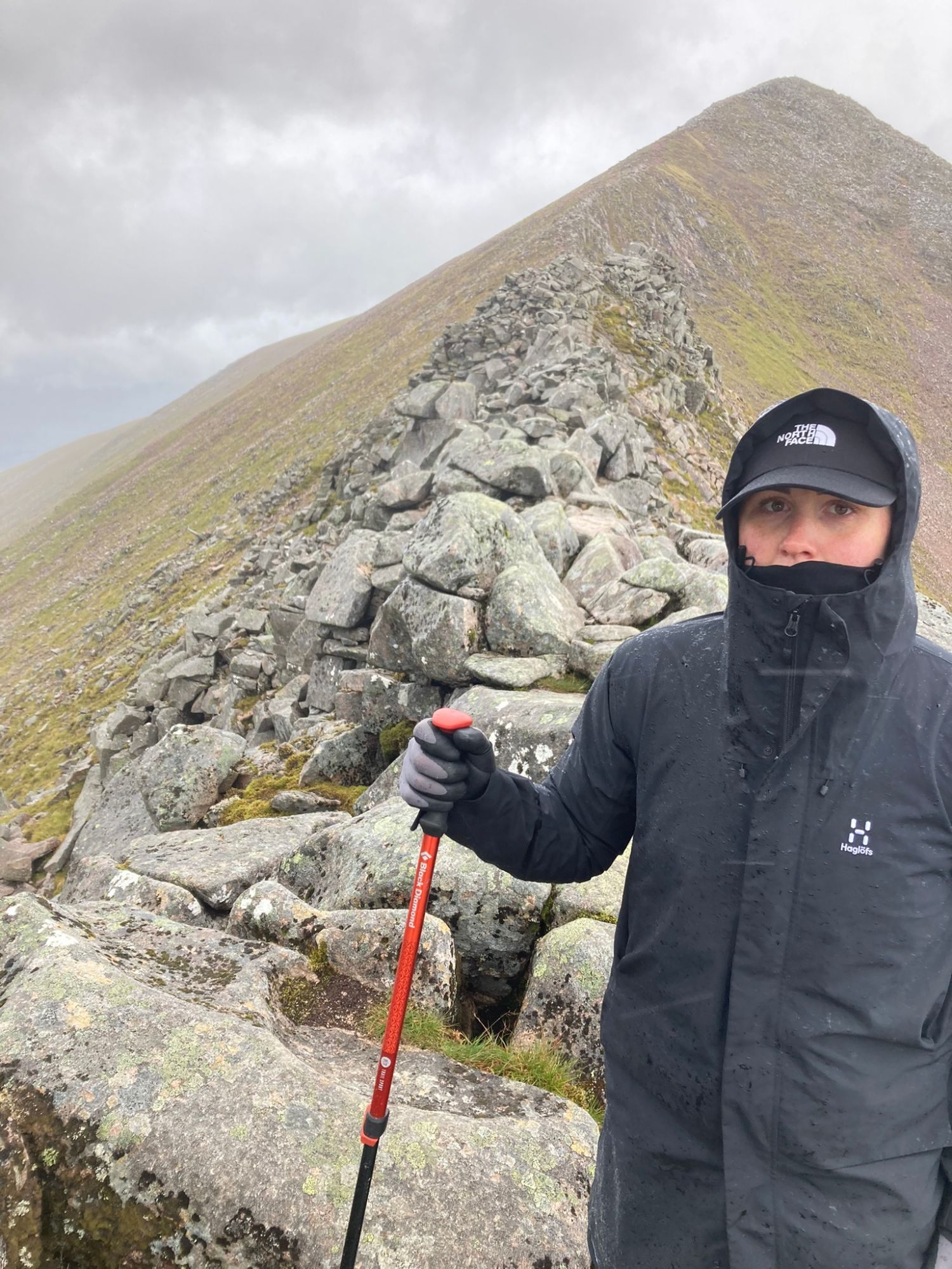 Kristine in a Haglöfs jacket and cap holding a red trekking pole on the boulder ridge, with a grassy slope behind