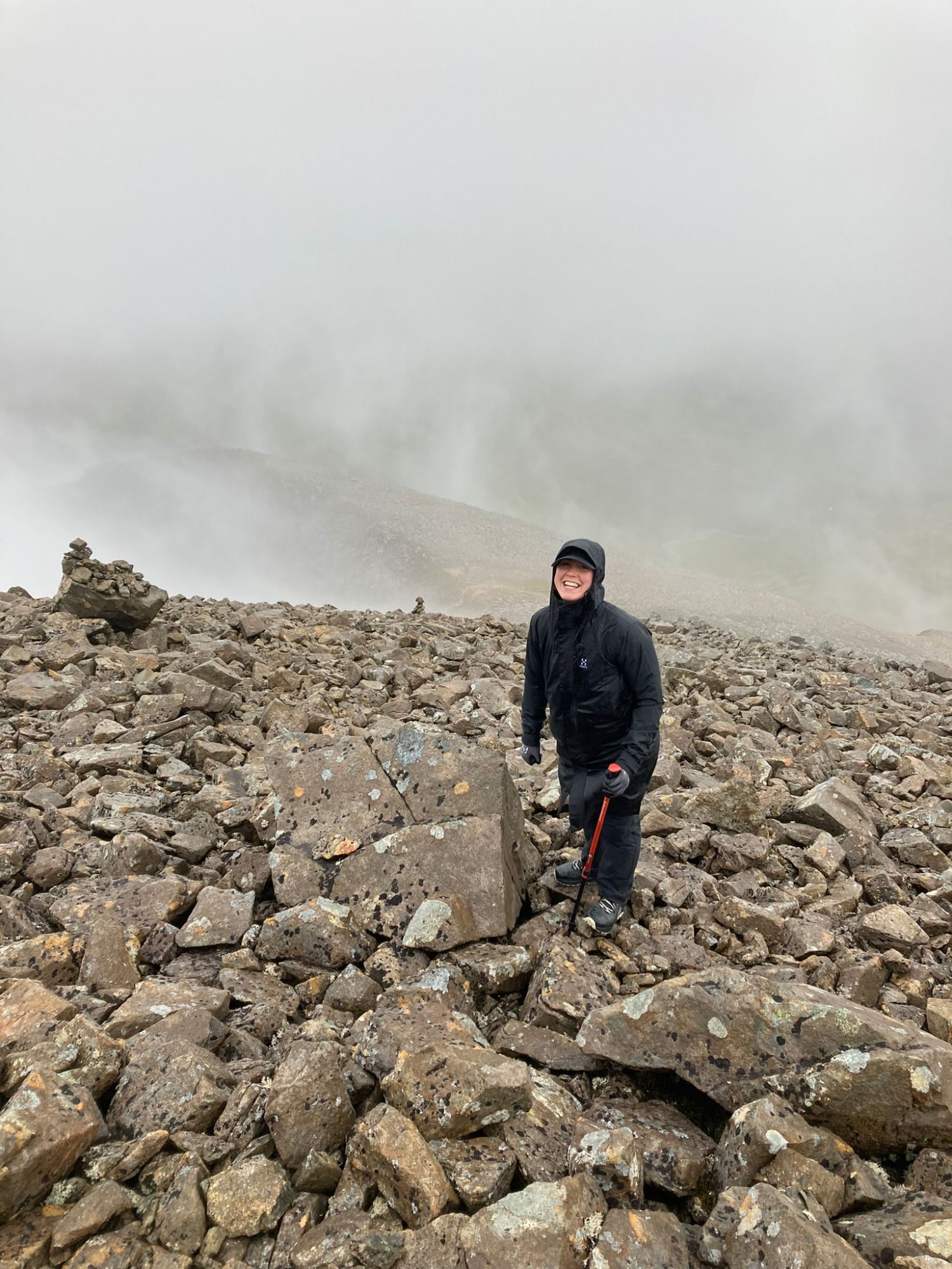 Kristine laughing on a boulder field near the top in thick cloud, with a cairn behind