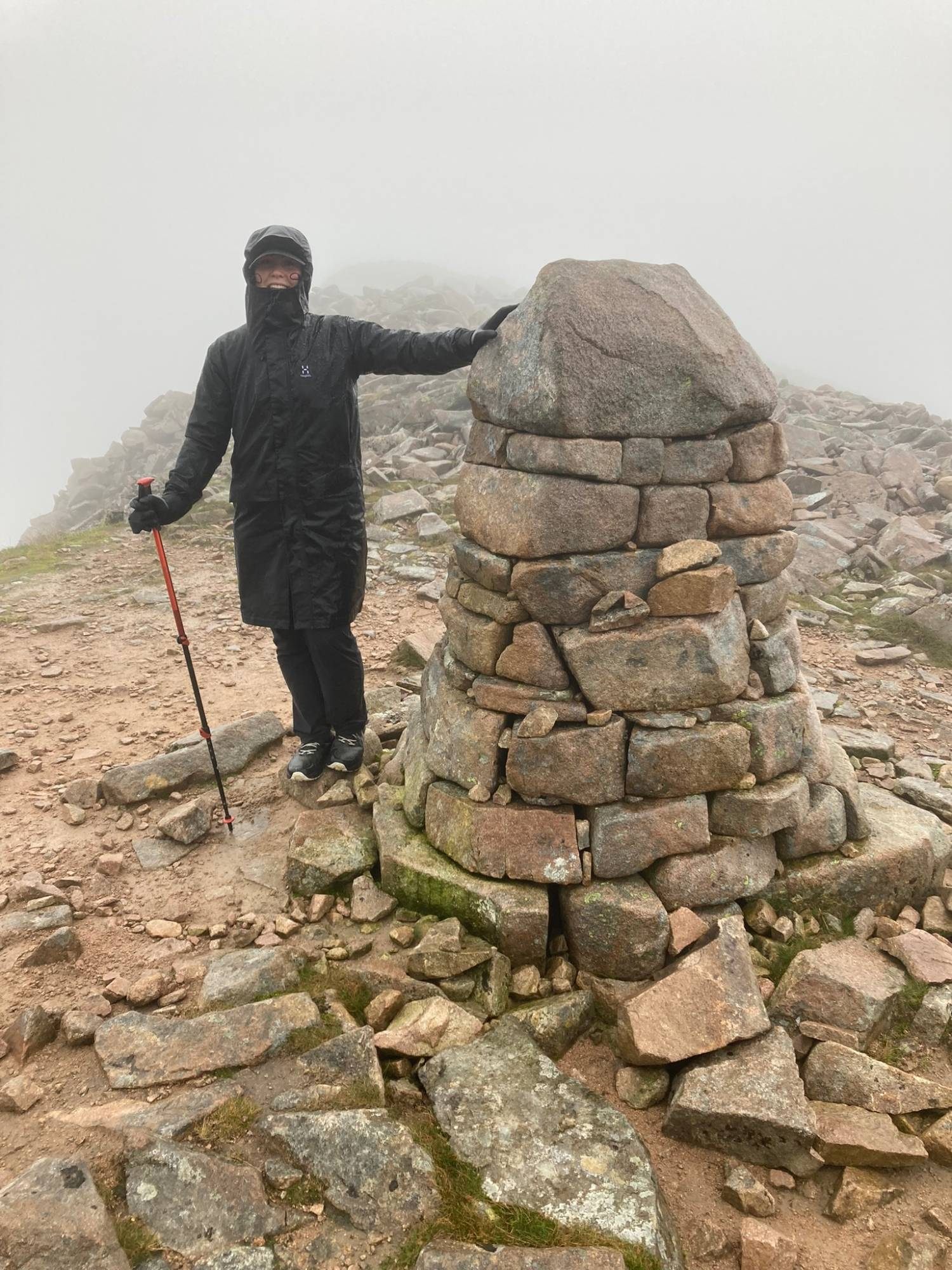 Kristine in a long rain jacket with a hand on a large stone cairn in thick cloud