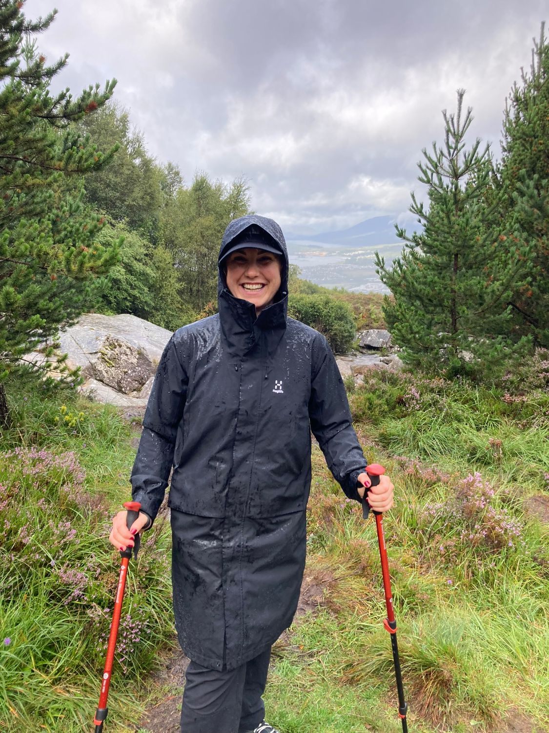 Kristine smiling in a wet rain jacket with two trekking poles on a forest path, with Fort William far below