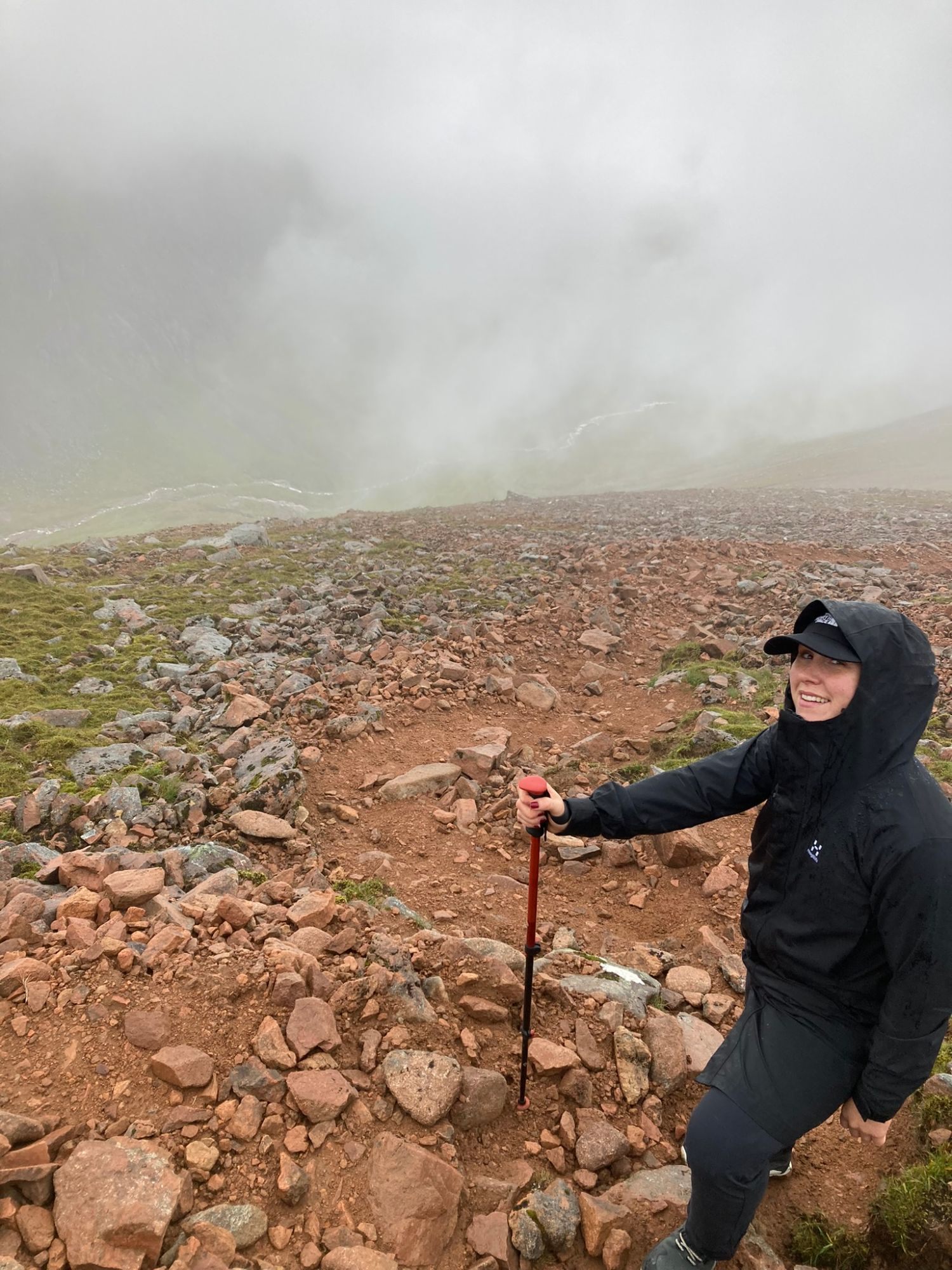 Kristine with a trekking pole on a steep path of red granite stones, with cloud filling the glen below
