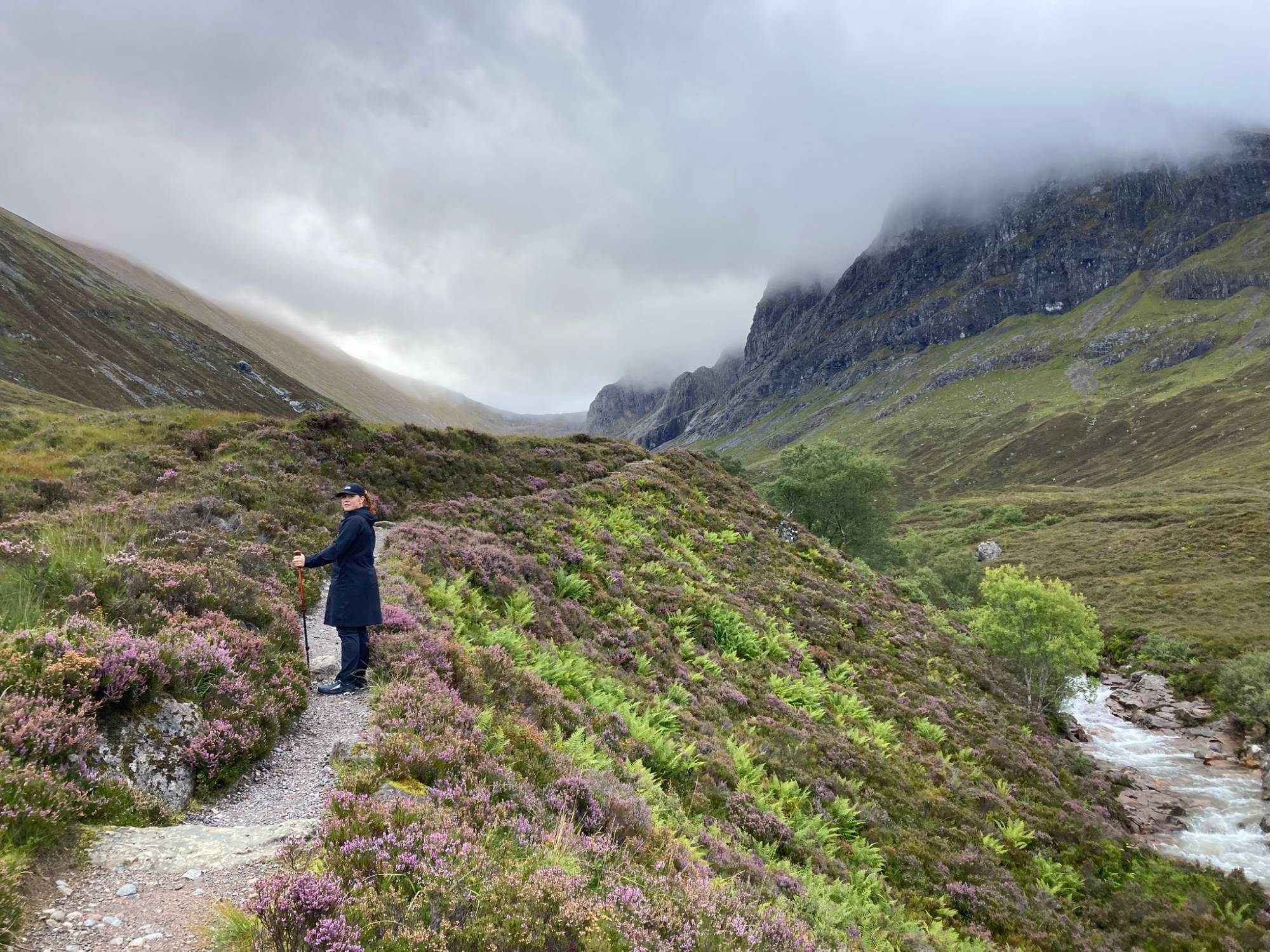 Kristine on a path through purple heather and ferns, with a rushing stream and the cloud-covered cliffs of the Ben Nevis north face