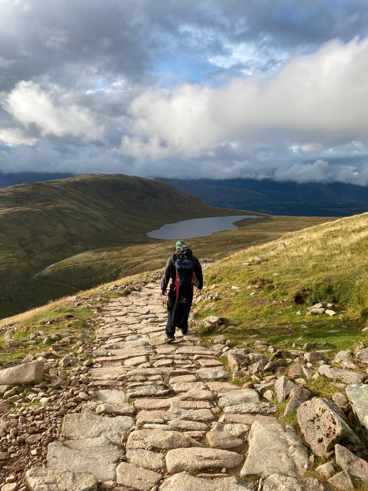 Leif walking down the stone-paved Mountain Track towards a lochan, with sunlit hills and clouds