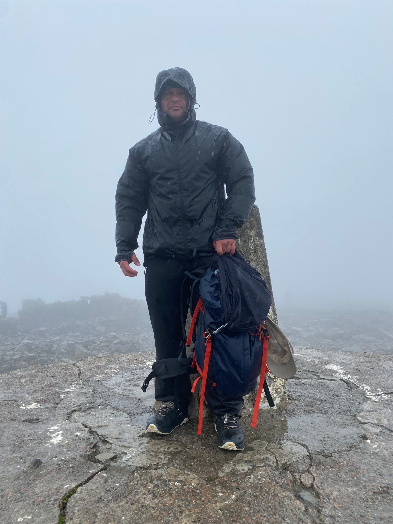 Leif in a wet rain jacket holding his backpack next to the trig pillar in thick cloud