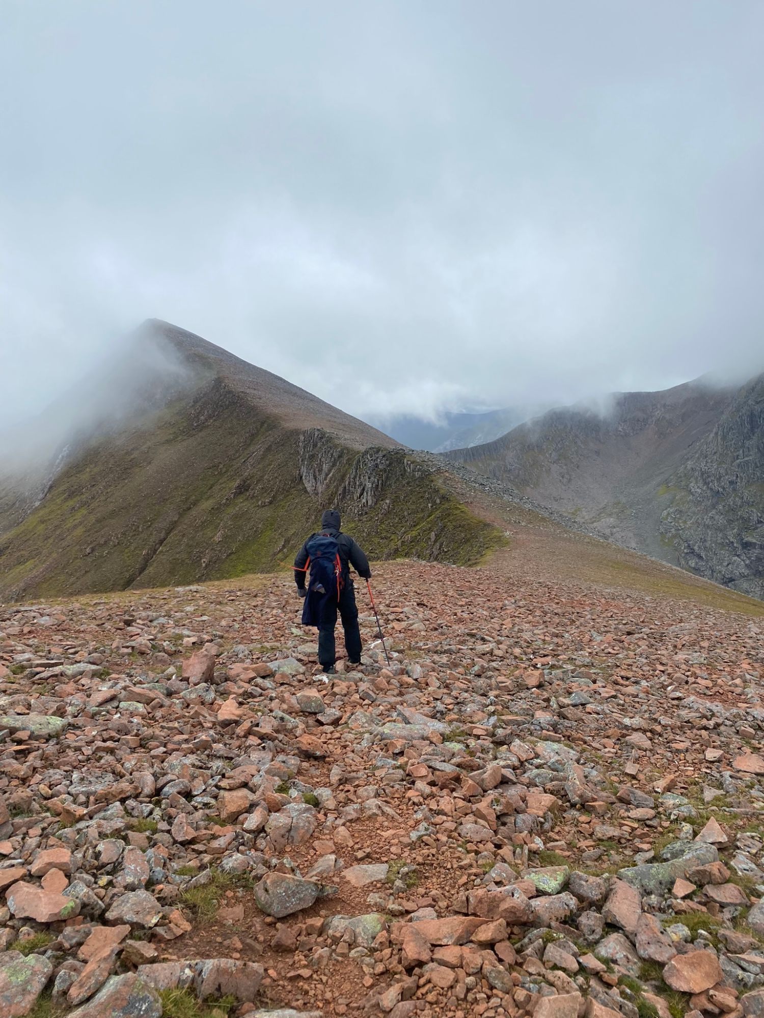 Leif with a trekking pole walking down red scree towards the narrow ridge of the CMD Arête, with the cliffs of Ben Nevis in cloud