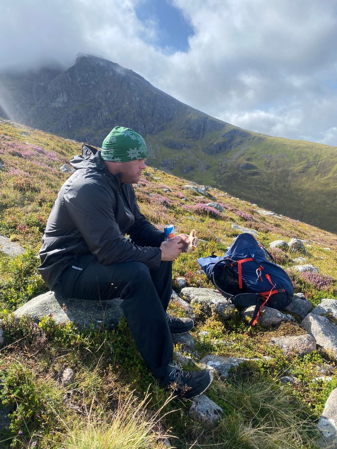 Leif in a green beanie eating a sandwich on a rock among heather, with sunlit cliffs behind