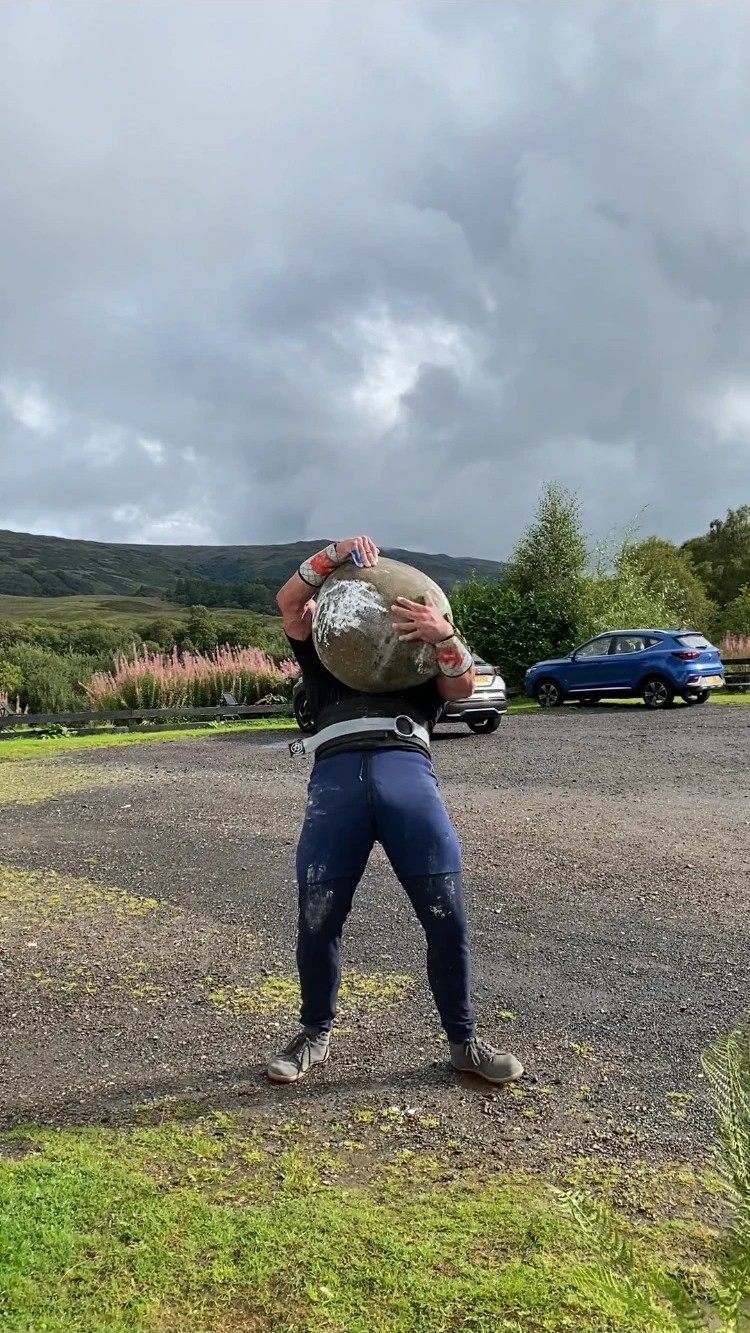 Leif holding the round MacGregor Stone at chest height in the hotel car park, with hills behind